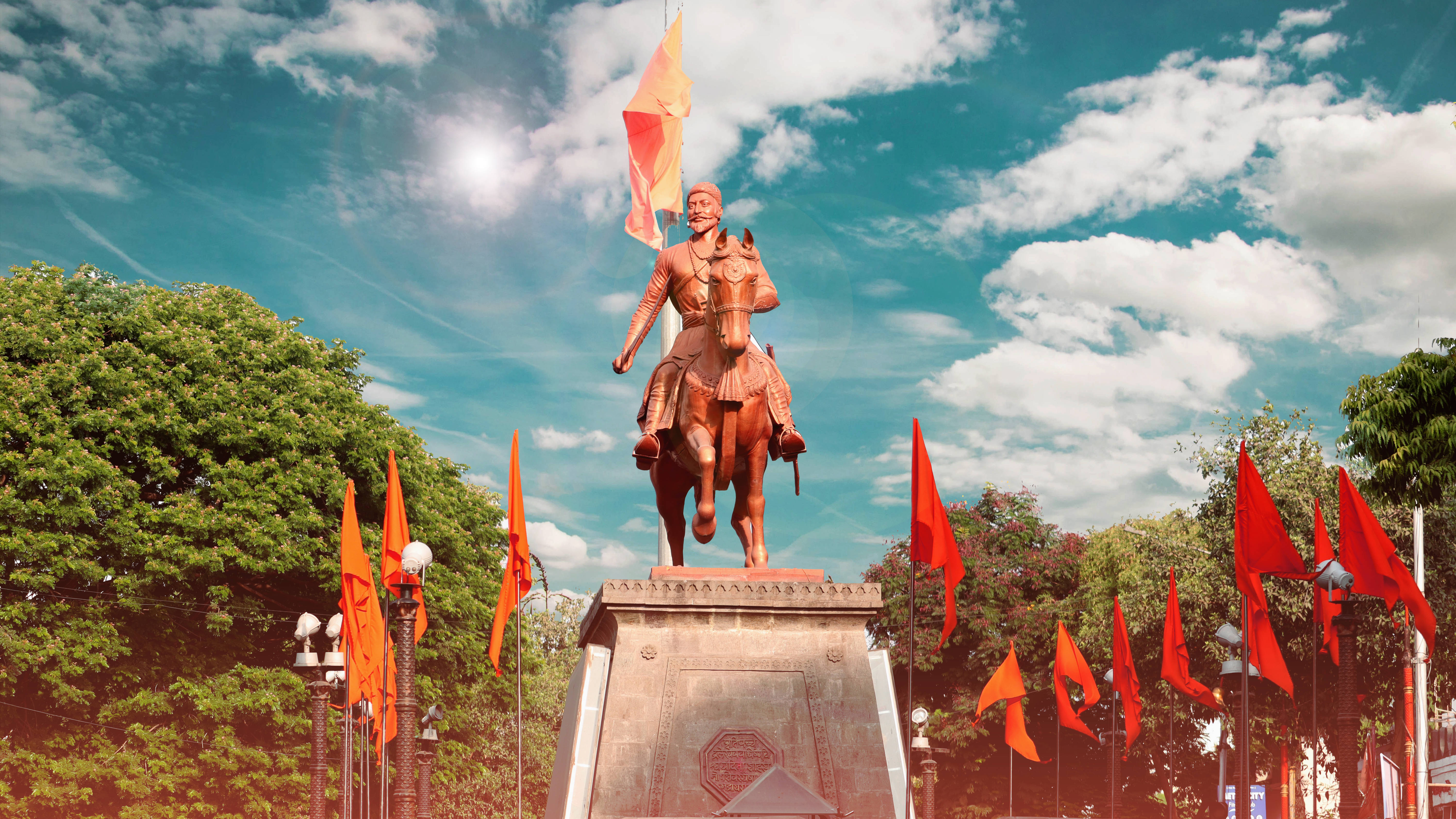 Indian 桌布 壁紙 — 一座青銅騎馬戰士雕像高舉旗幟，在公園中紅橙旗幟飄揚的明亮陽光下巍然矗立。