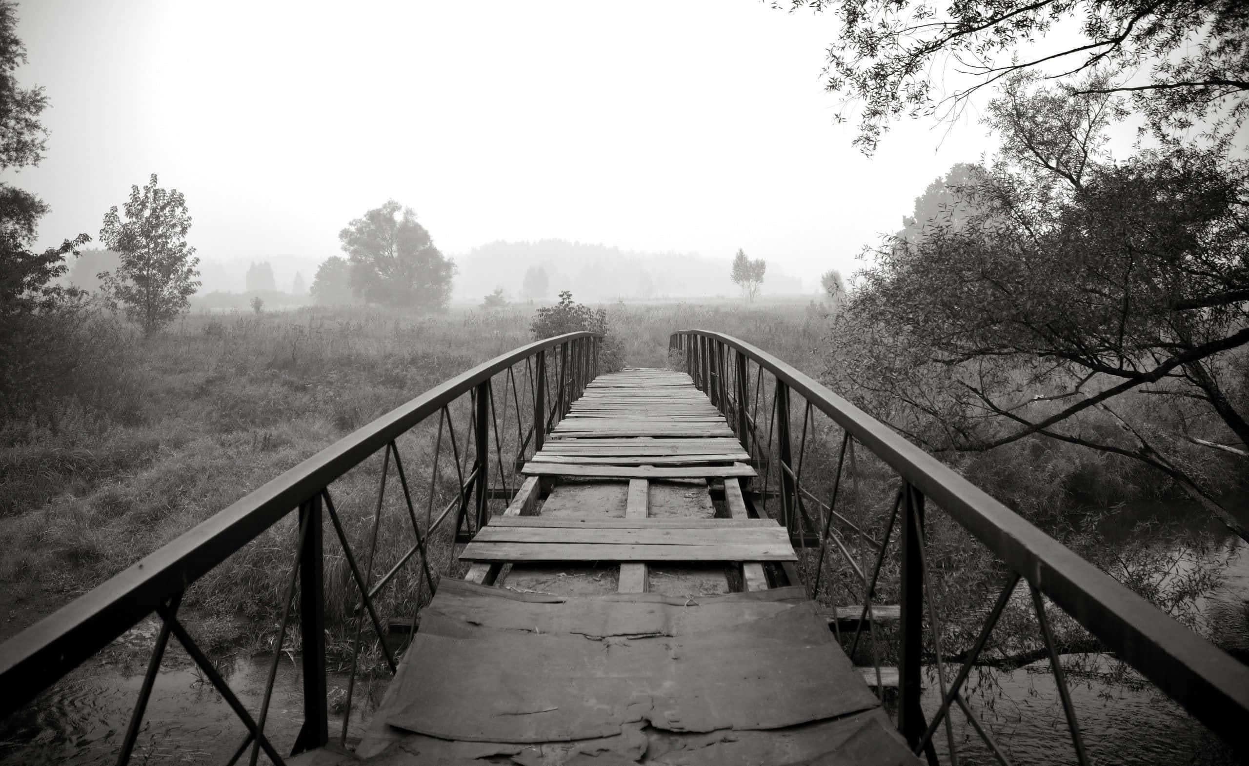 Black And White Landscape 桌布 壁紙 — 一座風化的木橋，帶有金屬欄杆，在陰天延伸進霧氣瀰漫的田野。