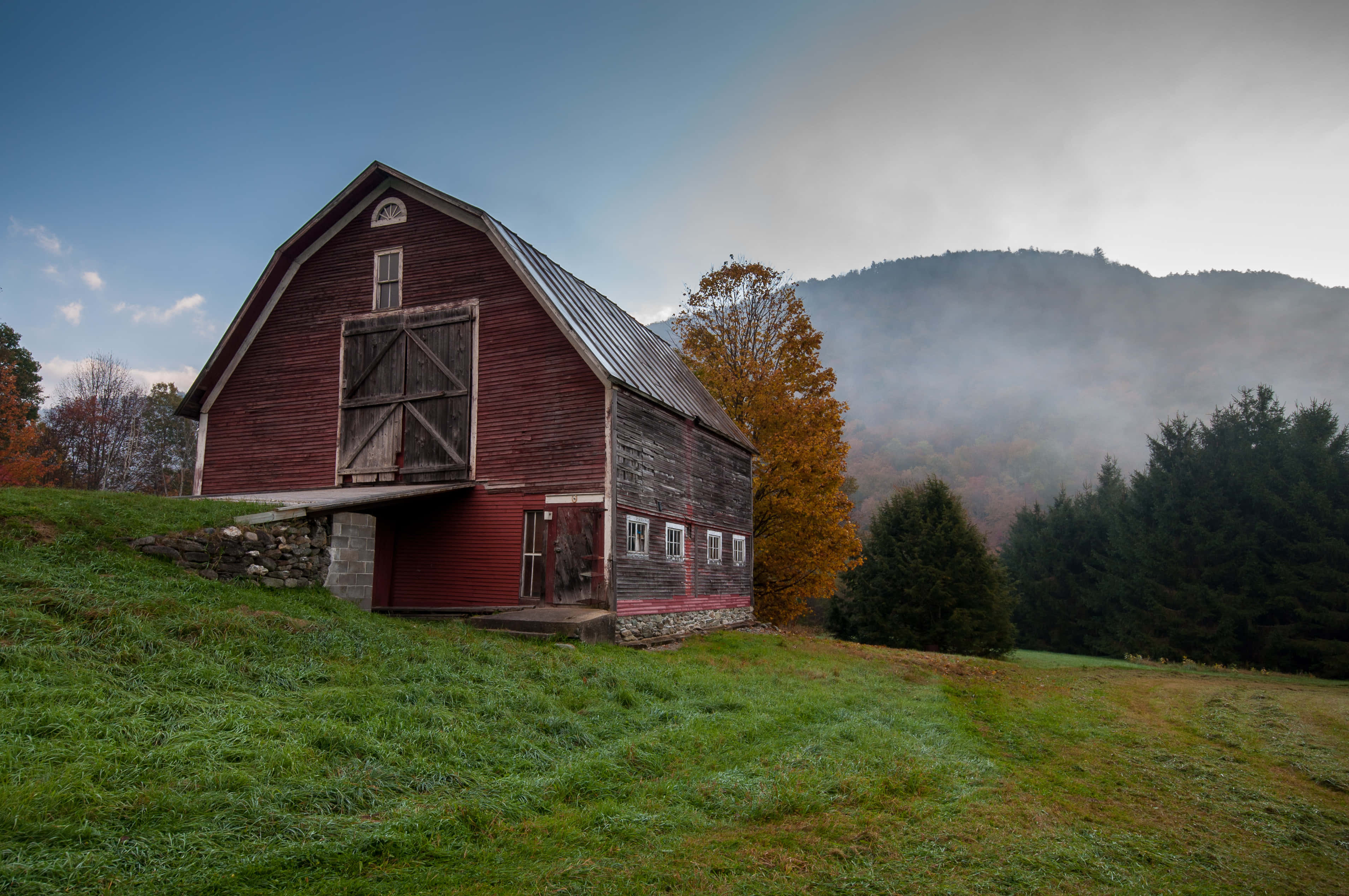 Fall Barn 桌布 壁紙 — 一座風化的紅色穀倉獨自矗立在長滿草的山丘上，背景有秋樹和霧濛濛的山巒。