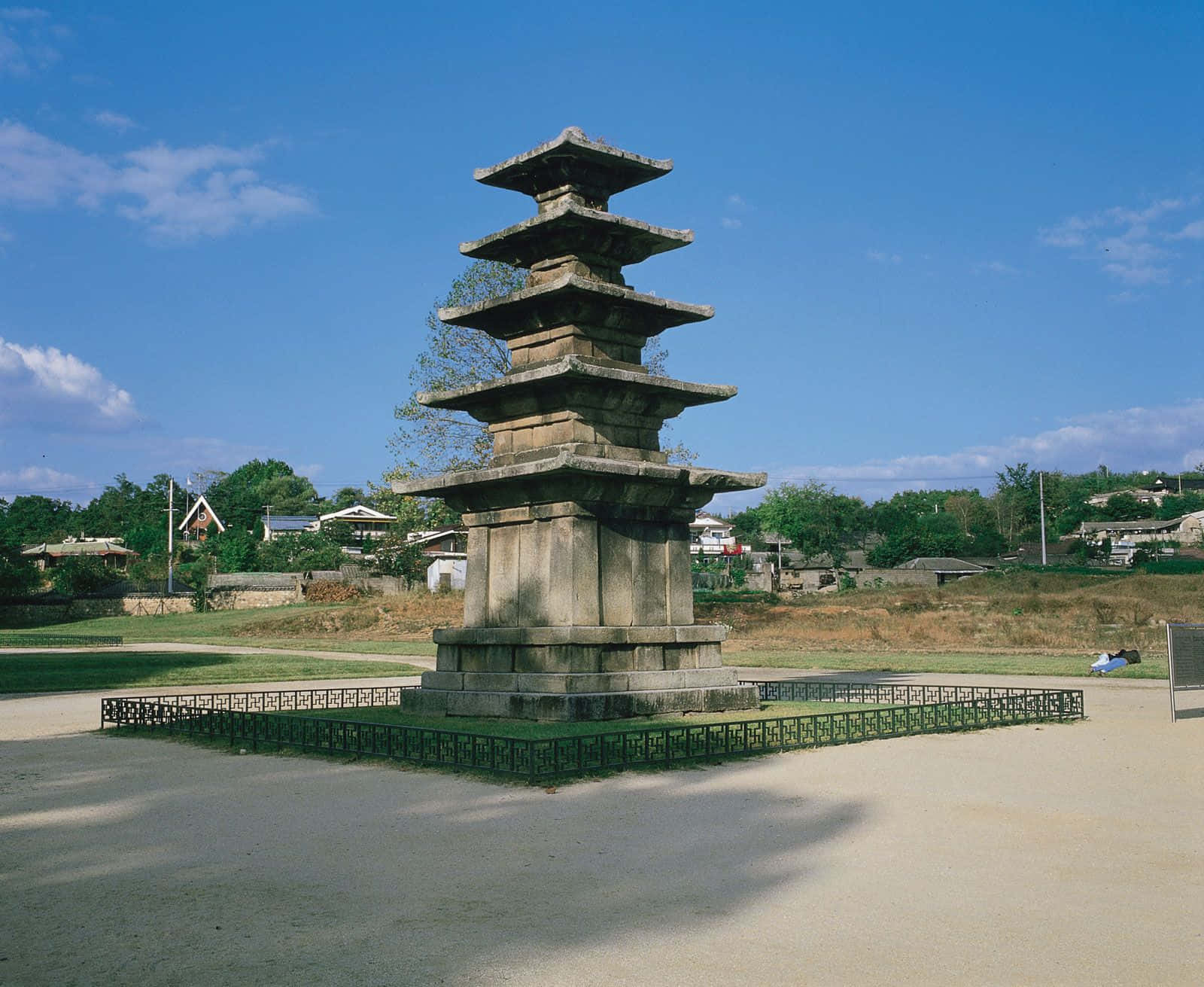 South Korea 桌布 壁紙 — 一座高大的古老石塔矗立在晴朗的藍天下，白雲點綴，周圍是綠草和遠處的村莊房屋。