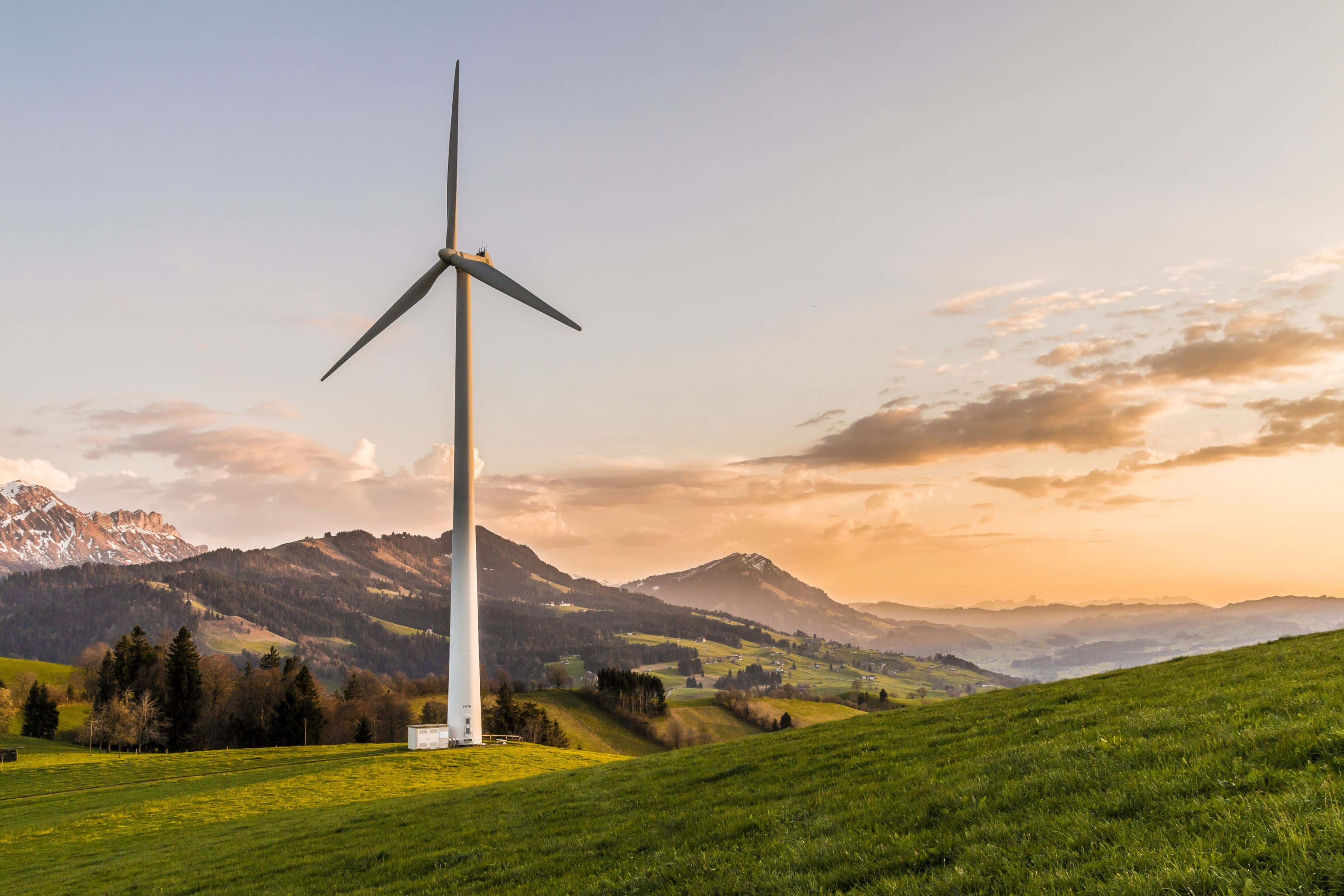 Renewable Energy 桌布 壁紙 — 一座高大的白色風力渦輪機矗立在夕陽下充滿活力的綠色山坡上，背景是白雪覆蓋的山脈。