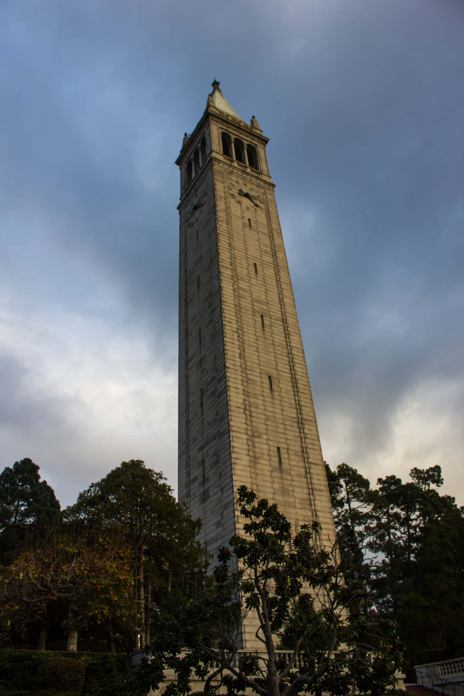 Ivory Tower 桌布 壁紙 — 一座高大的石造鐘樓聳立在戲劇性的多雲天空下，被深綠色的樹木環繞。