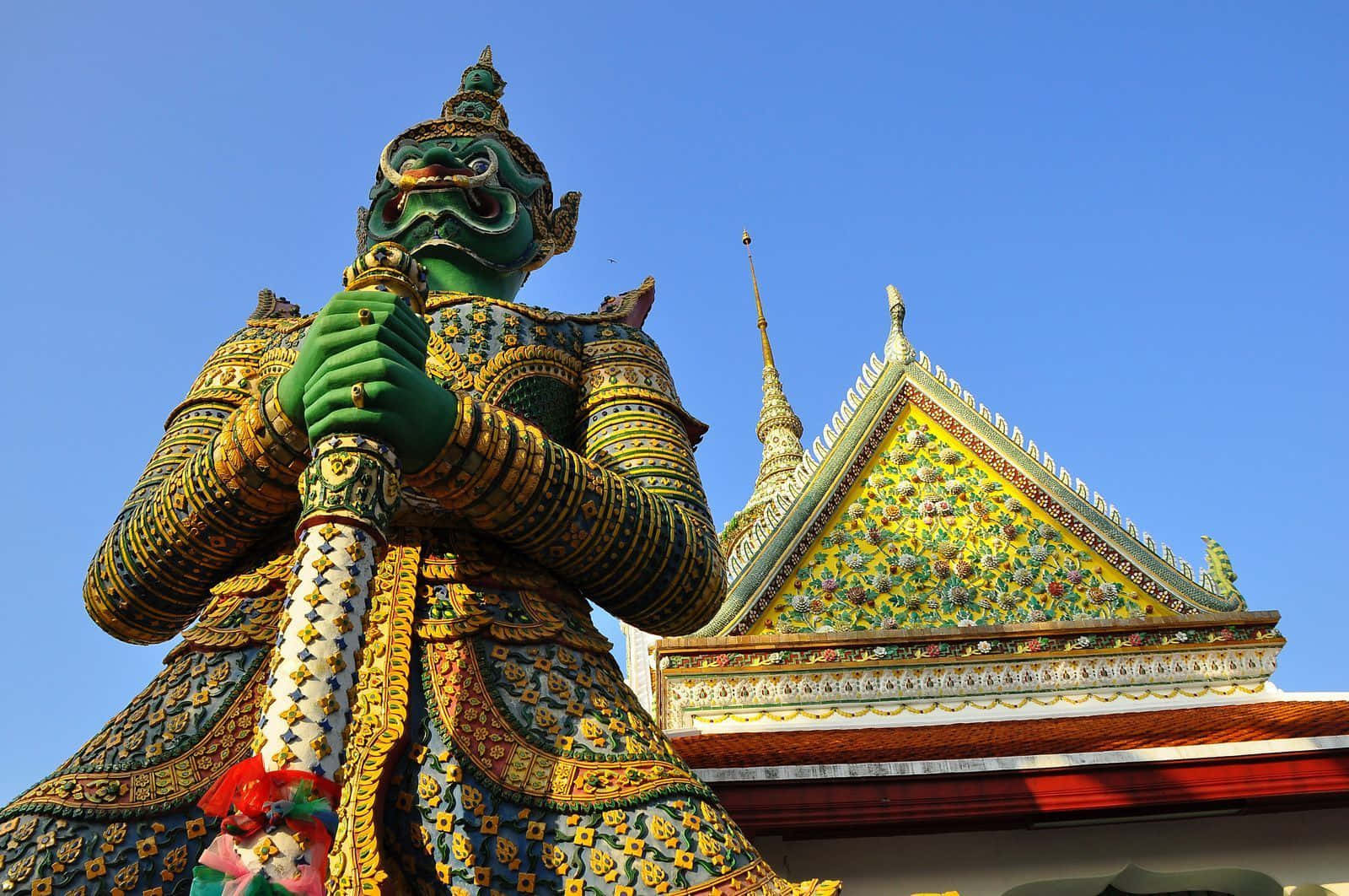 Thailand 桌布 壁紙 — 一座高大的綠色守護神雕像，身著華麗的金色盔甲，矗立在色彩鮮豔的泰國寺廟屋頂前，背景是清澈的藍天。