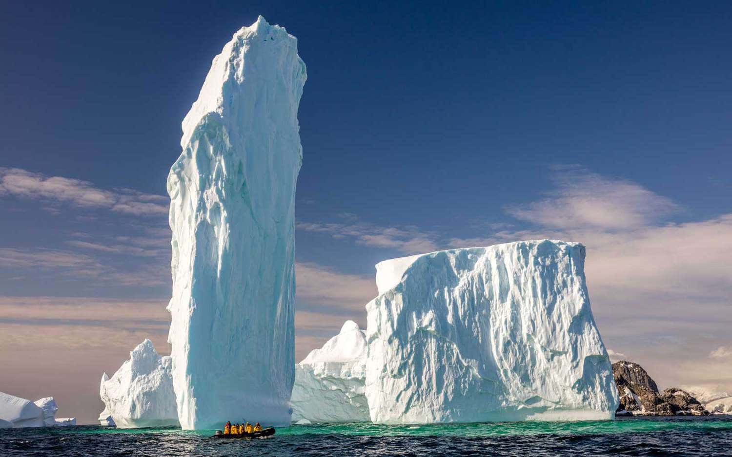 Antarctica 桌布 壁紙 — 一座高聳的冰山從碧綠的海水中雄偉地升起，在清澈的藍天下，附近有一艘小船。