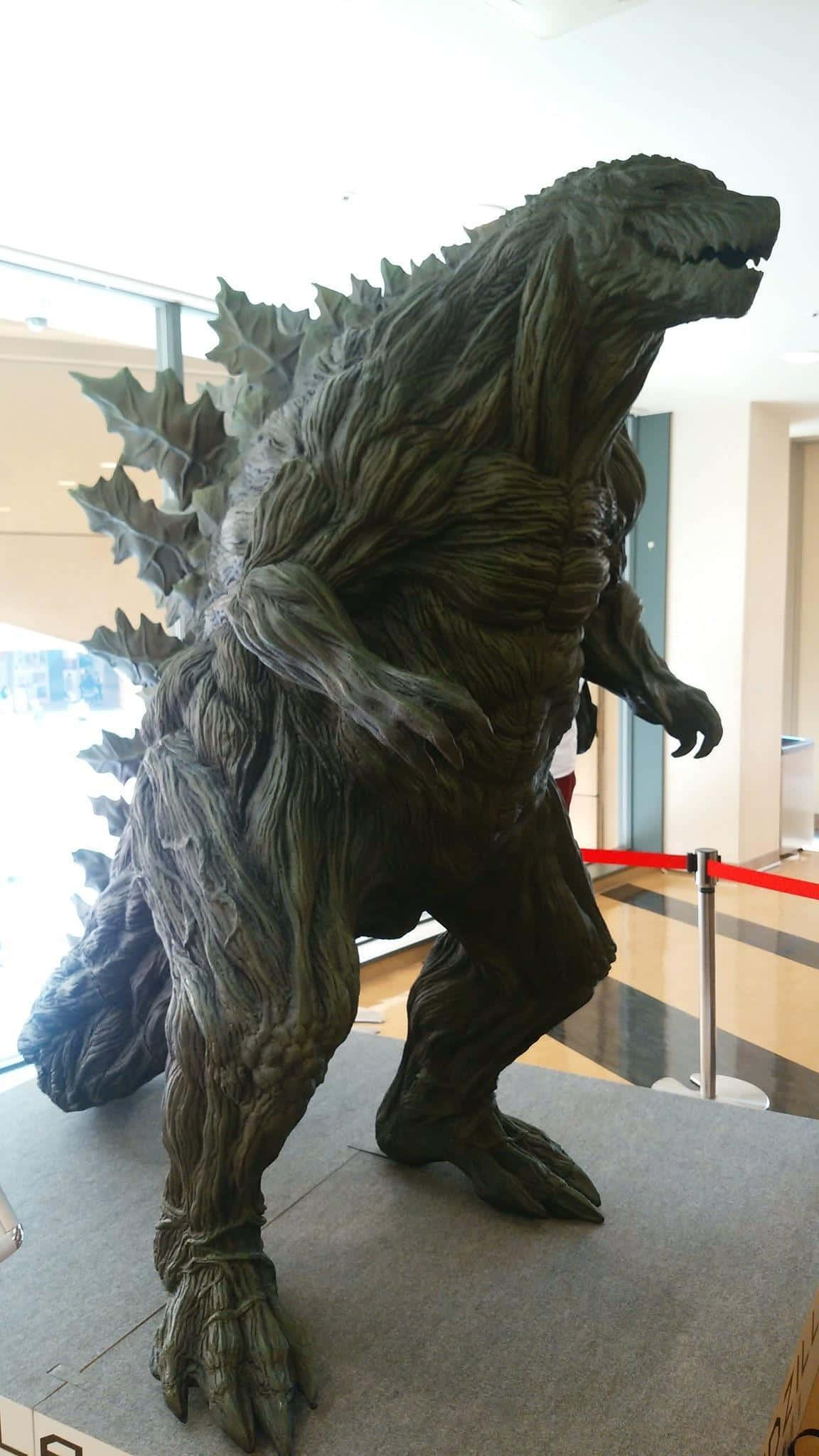 Godzilla Earth 桌布 壁紙 — 一座高聳的哥斯拉雕像，帶有紋理的綠褐色皮膚和尖刺背部，在現代室內大堂展出。
