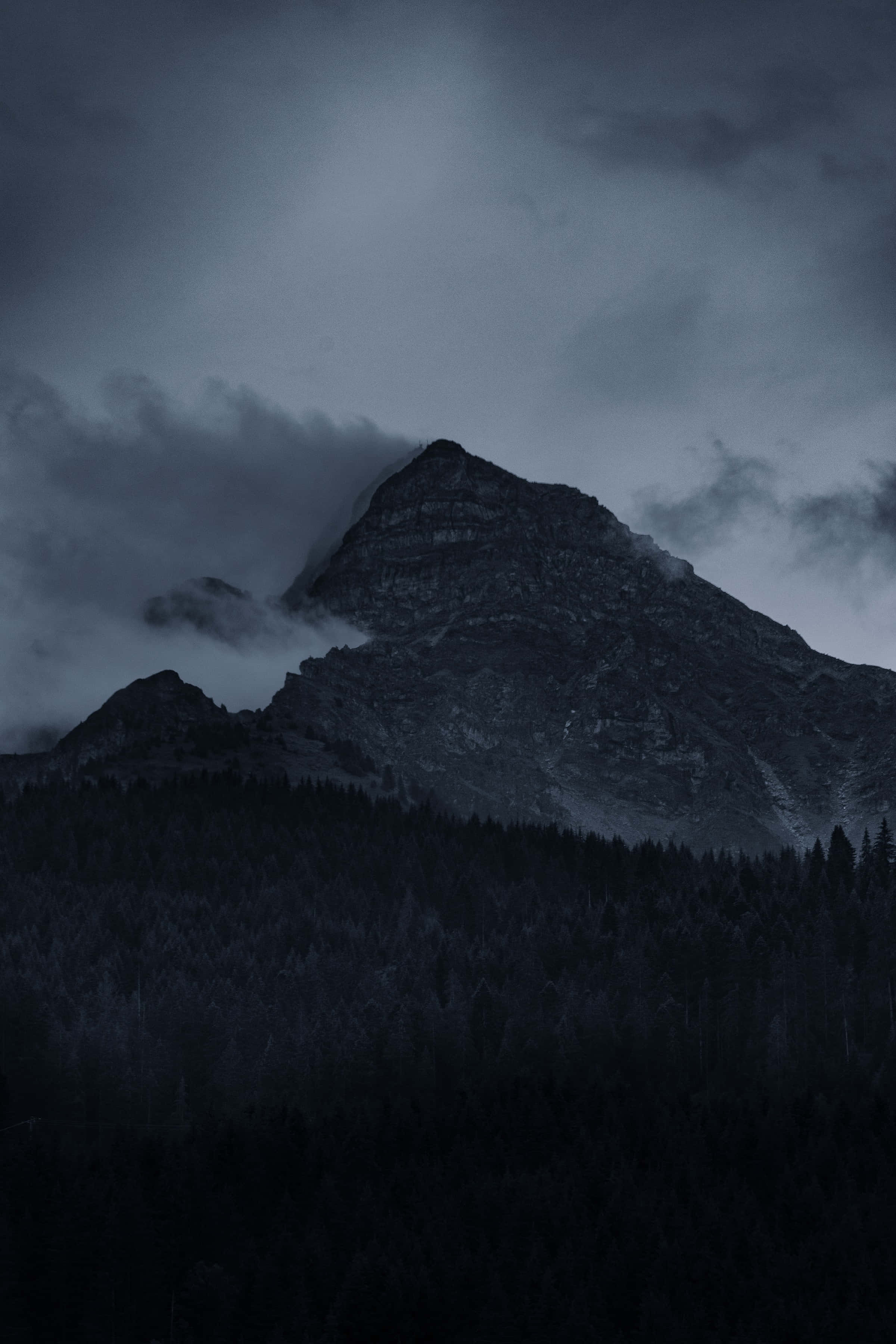 Dark Mountain 桌布 壁紙 — 一座高聳的山峰在黑暗的暮色天空下，穿過陰鬱的雲層，聳立於茂密的森林之上。