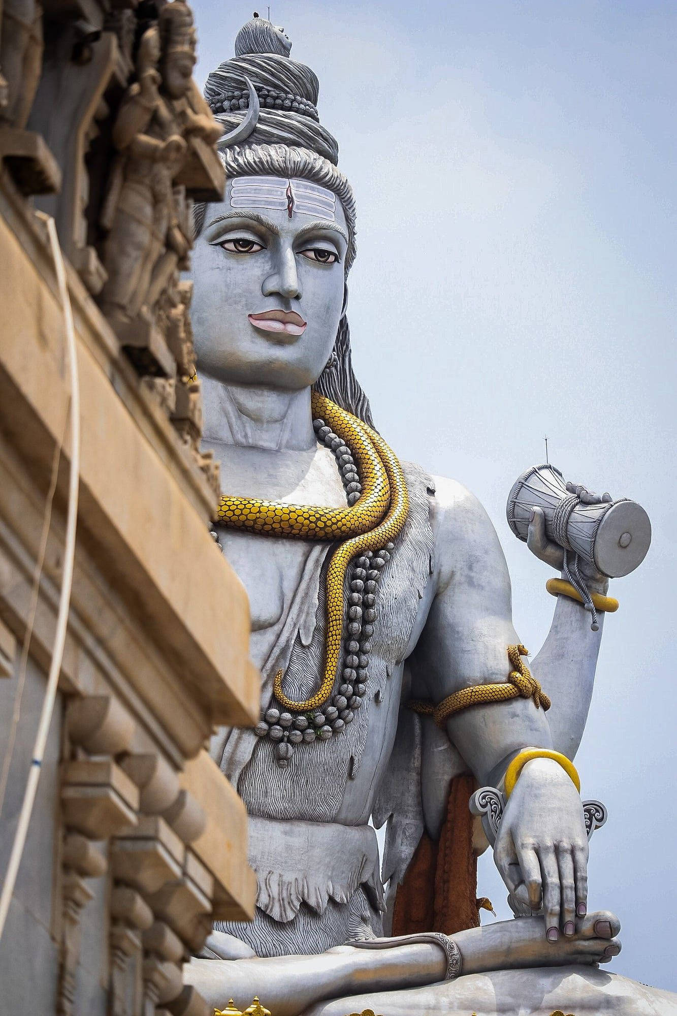 shiv shankar HD 桌布 壁紙 — 一座高聳的濕婆神石像，戴著金色蛇項鍊和達瑪魯鼓，背景是蒼白的天空。