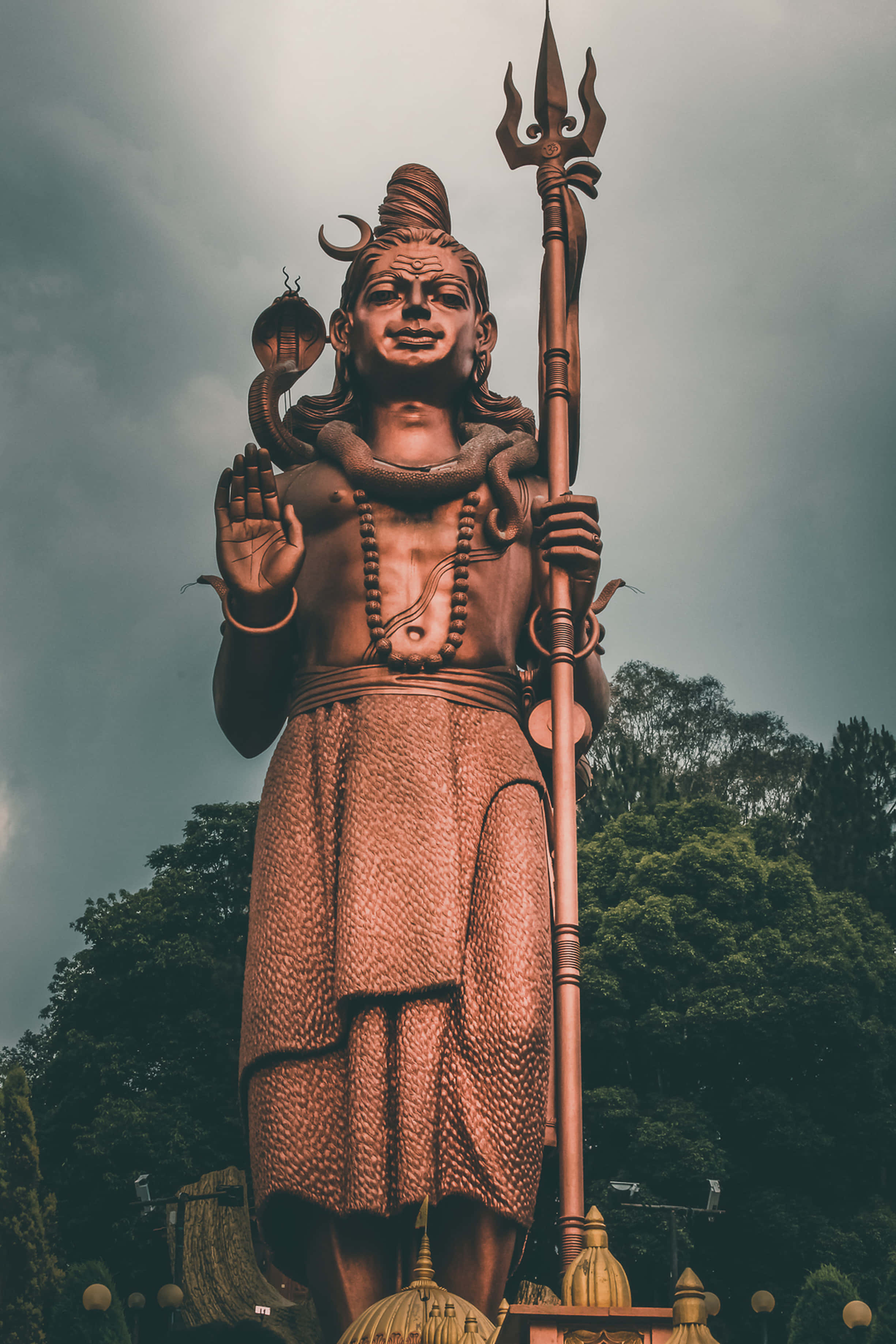 Mahadev 桌布 壁紙 — 一座高聳的濕婆青銅雕像，在暴風雨的天空下，背後是茂密的綠樹，威嚴矗立。