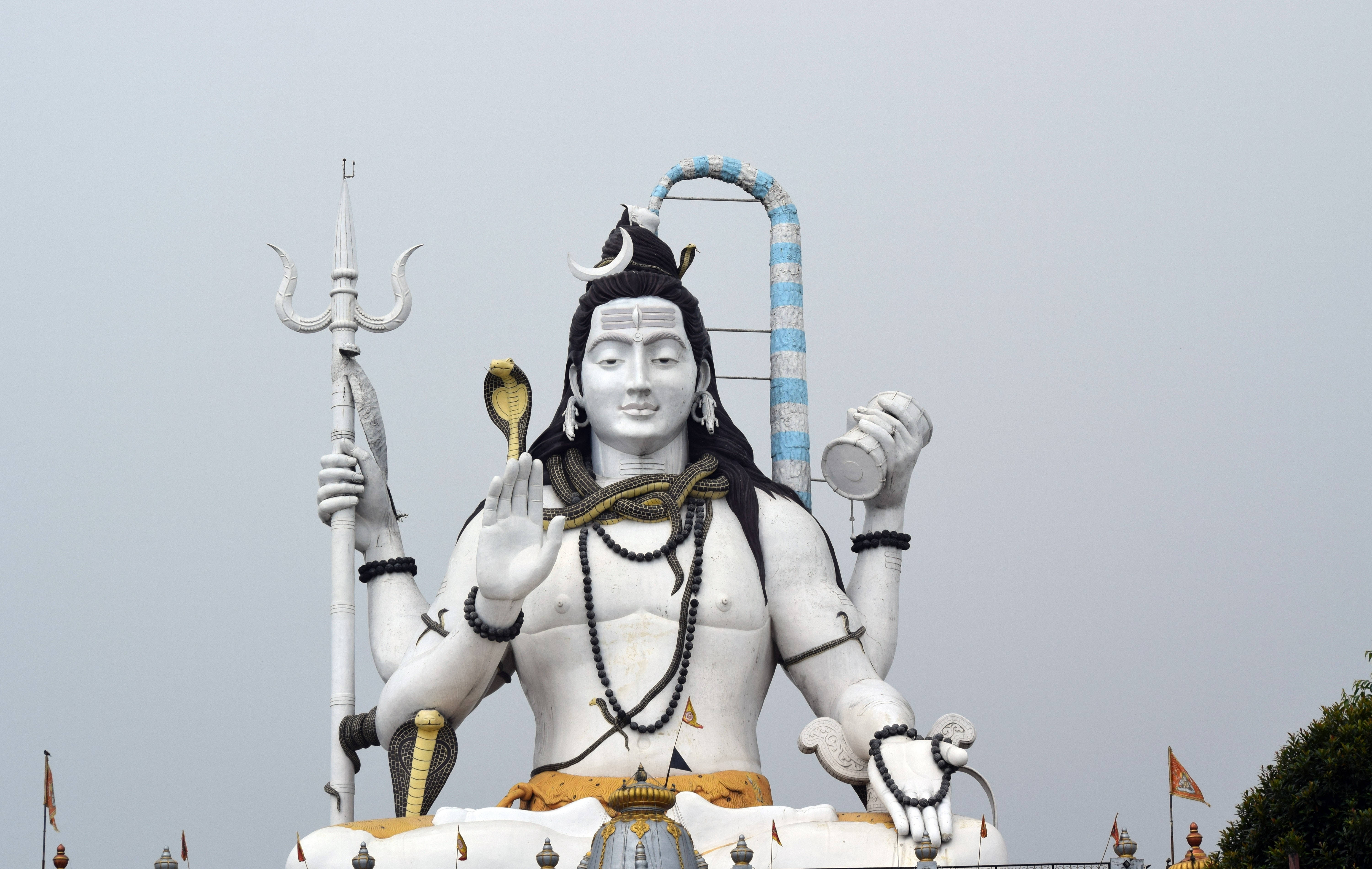 Lord Shiva 桌布 壁紙 — 一座高聳的白色濕婆神像，多臂手持三叉戟、蛇和鼓，背景是灰色天空。