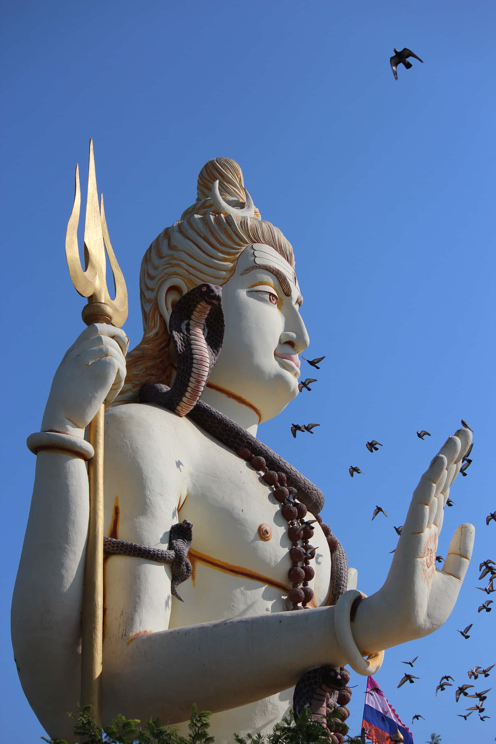 Mahadev 桌布 壁紙 — 一座高聳的白色濕婆神像，手持金色三叉戟，頸上纏繞著眼鏡蛇，背景是清澈的藍天，鳥兒在周圍飛翔。