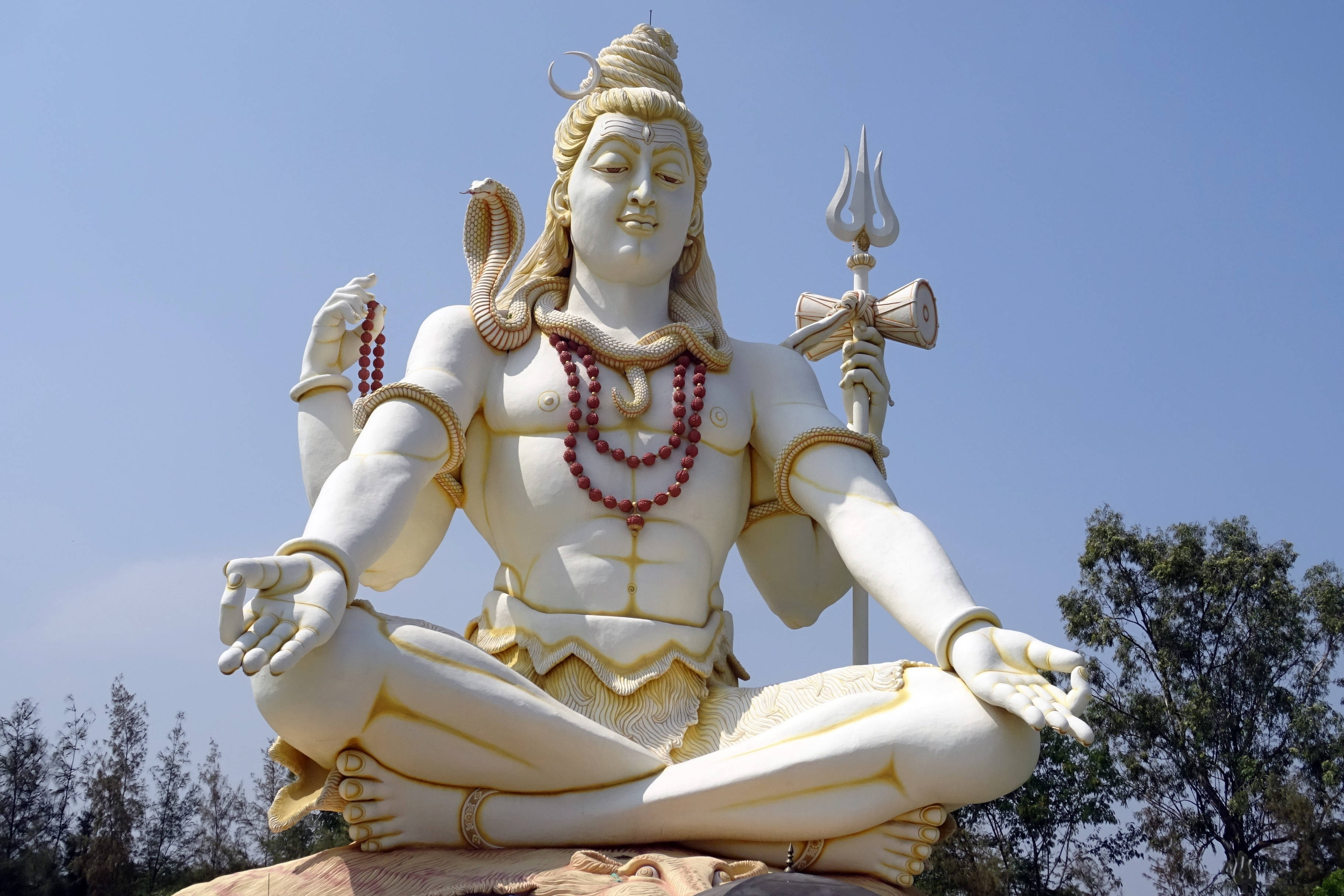 Lord Shiva 桌布 壁紙 — 一座高聳的白色石雕濕婆神像，呈冥想姿態，手持三叉戟和蛇，背景是清澈的藍天和下方的樹木。