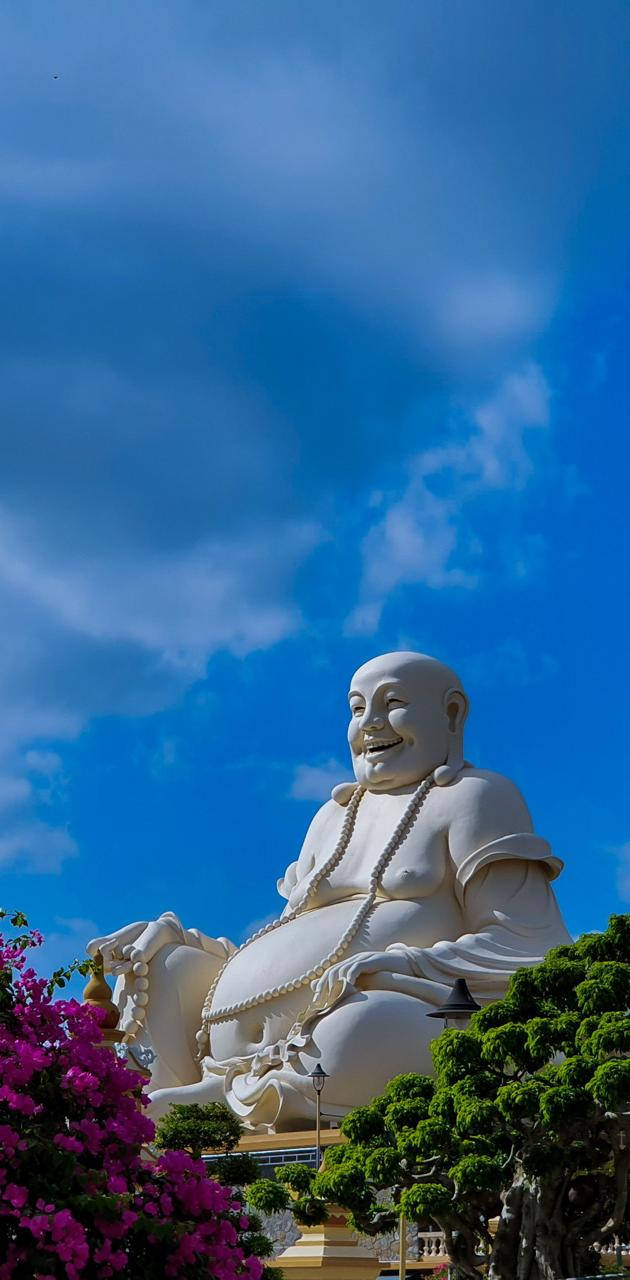 Statue Aesthetic 桌布 壁紙 — 一座高聳的白色笑佛雕像在藍天白雲和下方色彩繽紛的花朵襯托下，寧靜地微笑。