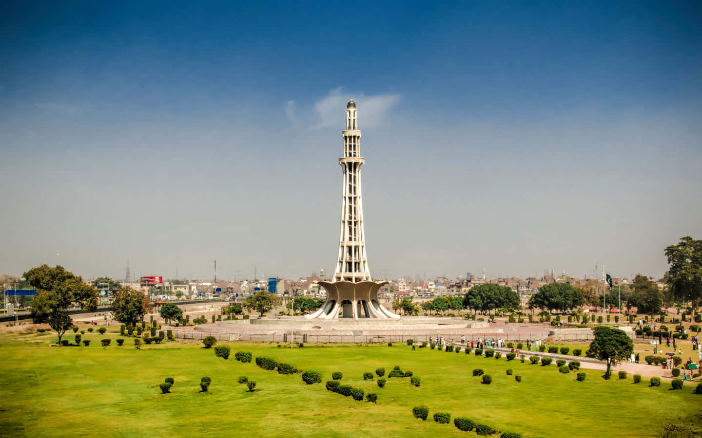 Pakistan 桌布 壁紙 — 一座高聳的白色紀念碑在晴朗的藍天下，矗立於廣闊的綠色公園中，遠處可見城市建築。