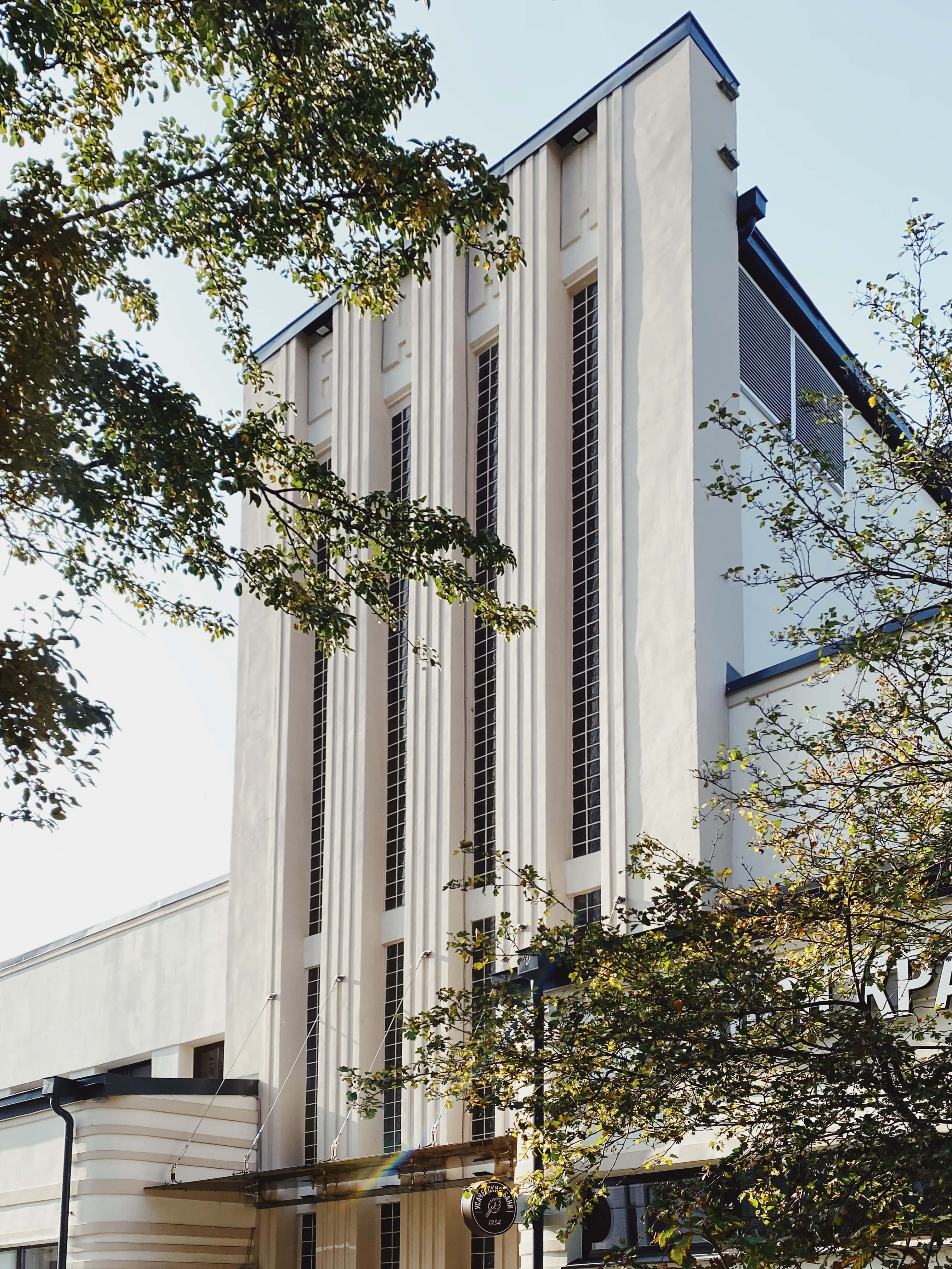Constructivism 桌布 壁紙 — 一座高聳的白色裝飾藝術風格建築，帶有垂直線條和藍色飾邊，在晴朗的天空下矗立在茂密的綠樹之後。
