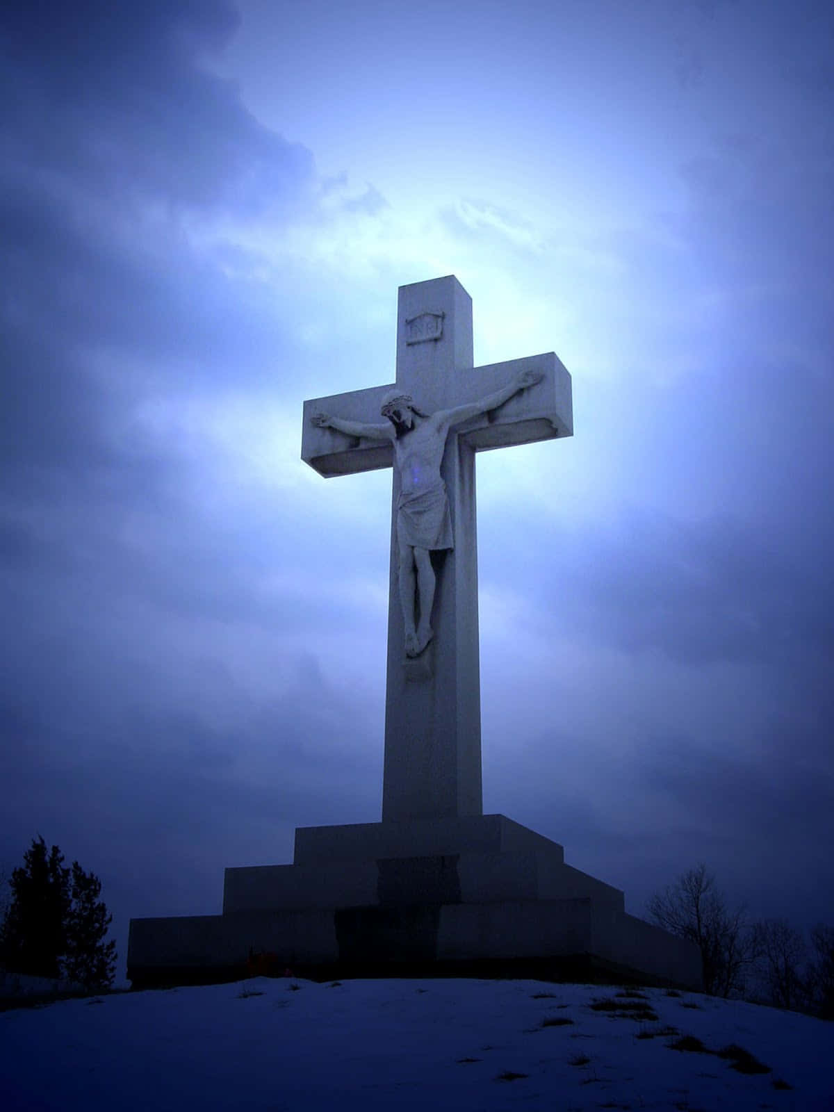 Cool Cross 桌布 壁紙 — 一座高聳的石製十字架雕像，在積雪的小山丘上，於陰沉的暮色天空下莊嚴地矗立。