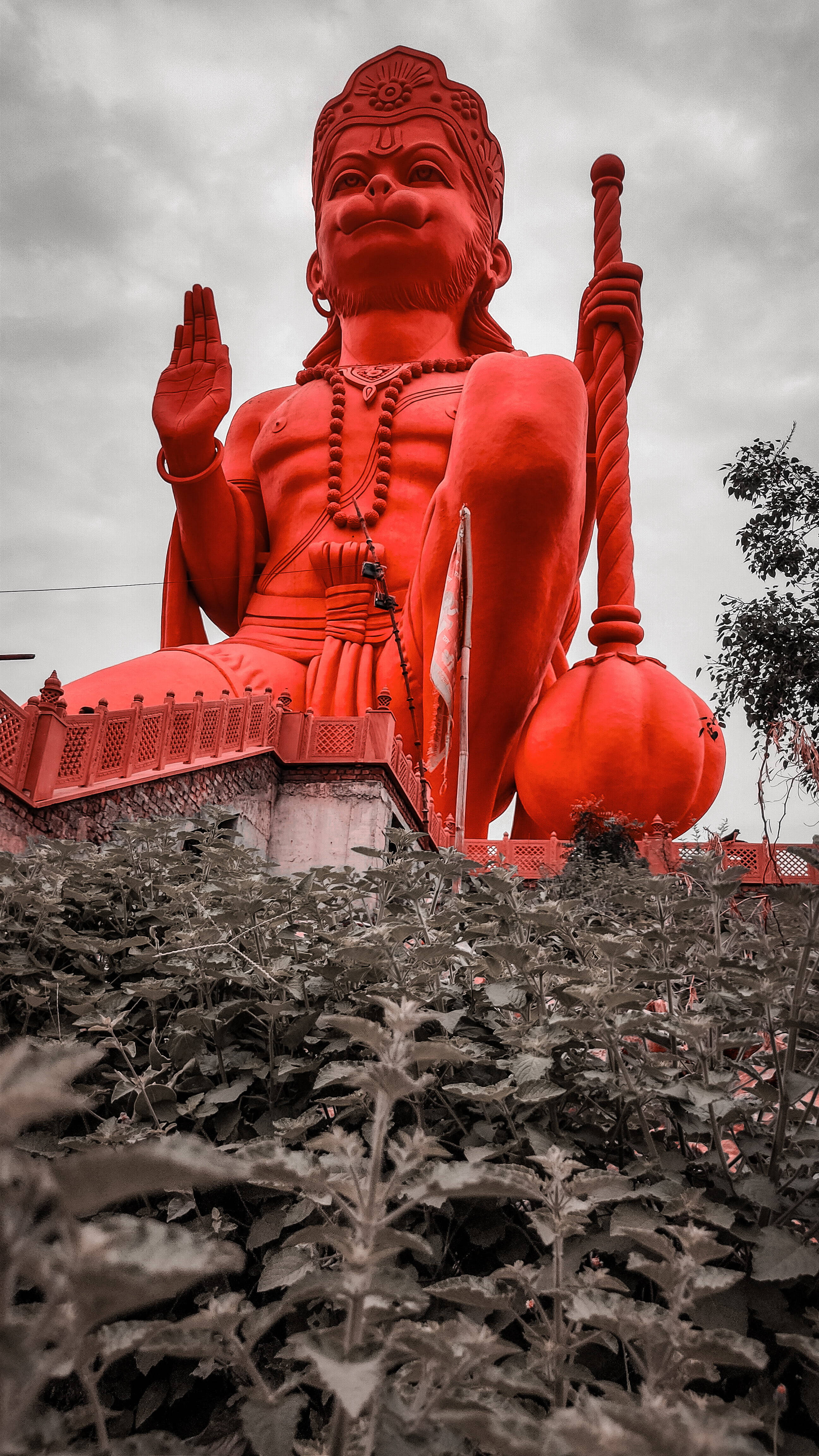 lord hanuman 桌布 壁紙 — 一座高聳的紅色哈努曼雕像在陰天天空下，寧靜地坐在被茂密綠色植物環繞的平台上。