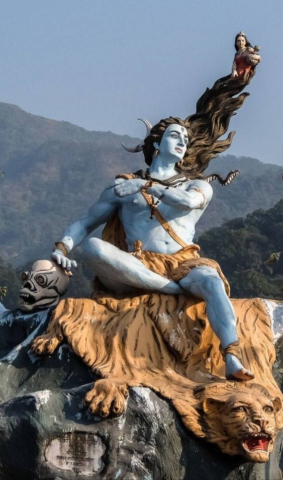 shiv shankar HD 桌布 壁紙 — 一座高聳的藍皮膚濕婆雕像，頭髮飄逸，身披虎皮，手持一個頭骨和一個小雕像，以山脈為背景。
