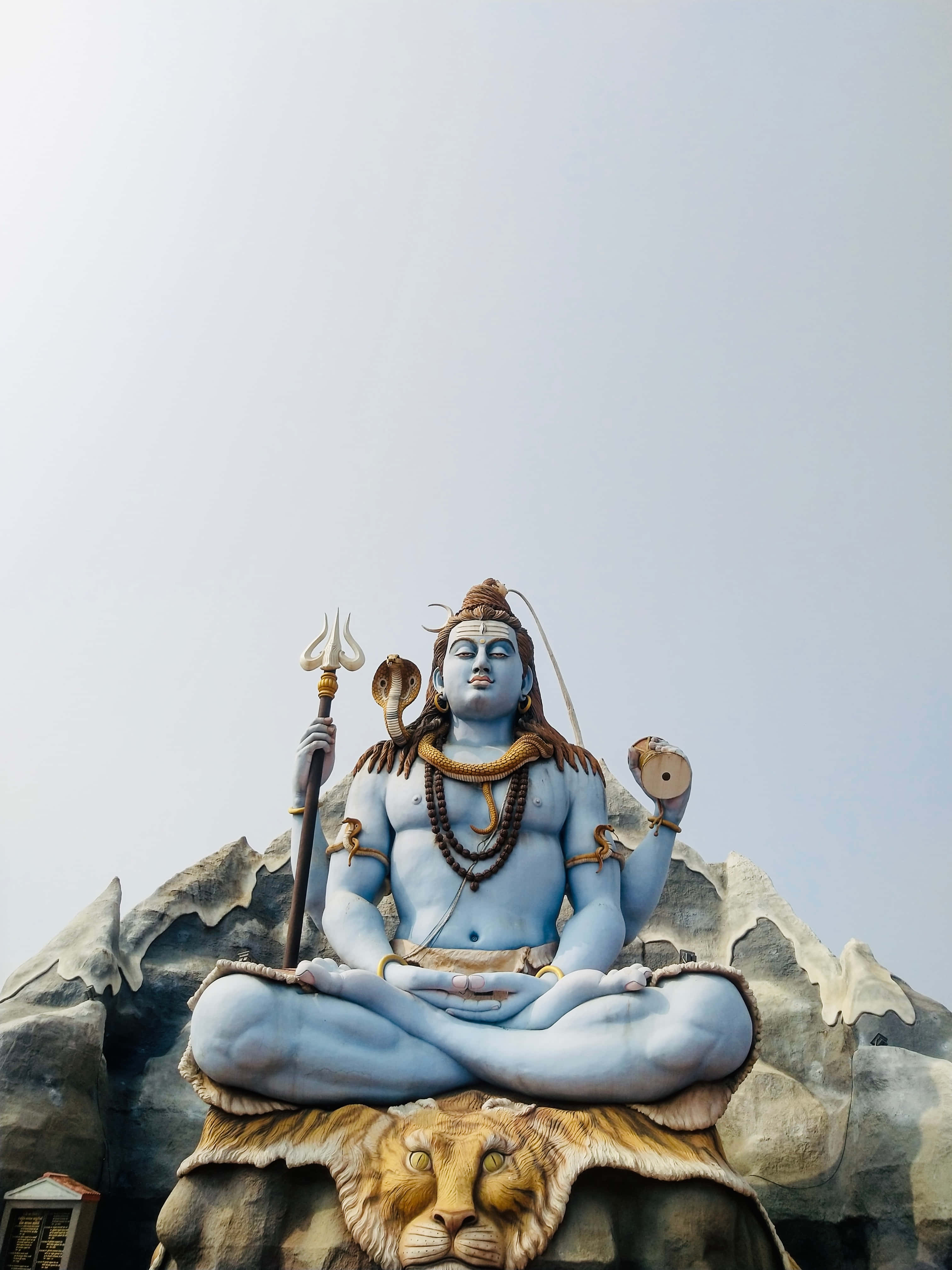 Mahadev 桌布 壁紙 — 一座高聳的藍色濕婆神像在獅子上冥想，手持三叉戟和達瑪魯，背景是蒼白的天空。