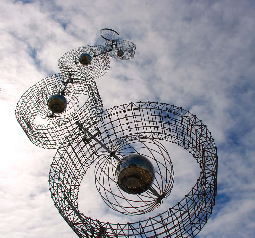 Kinetic Art 桌布 壁紙 — 一座高聳的金屬雕塑，帶有螺旋線框和反光球體，矗立在部分多雲的藍天下。