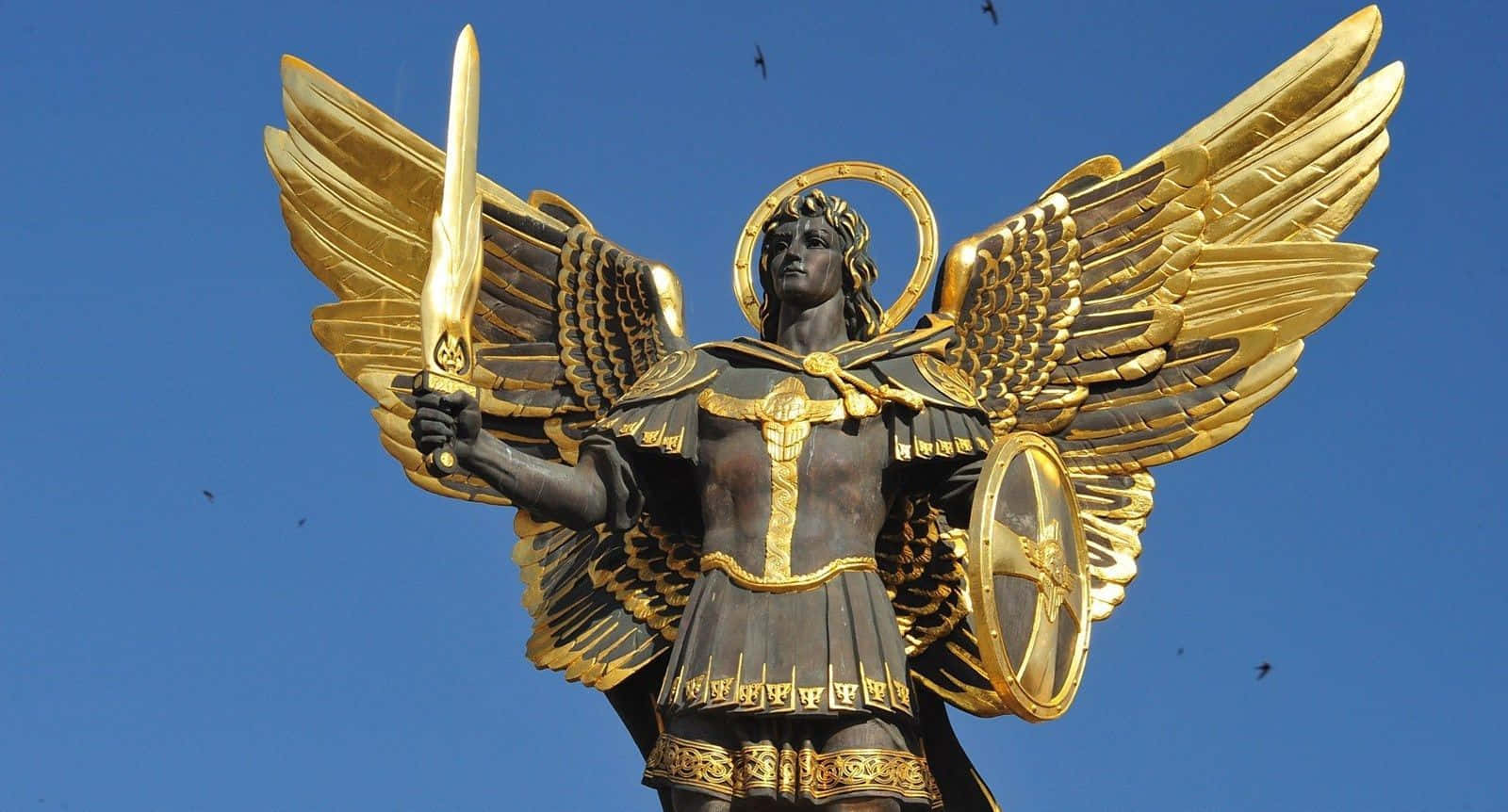 Saint Michael 桌布 壁紙 — 一座高聳的金翼天使雕像，身穿盔甲，手持劍與盾，背景是晴朗的藍天，有鳥兒飛翔。