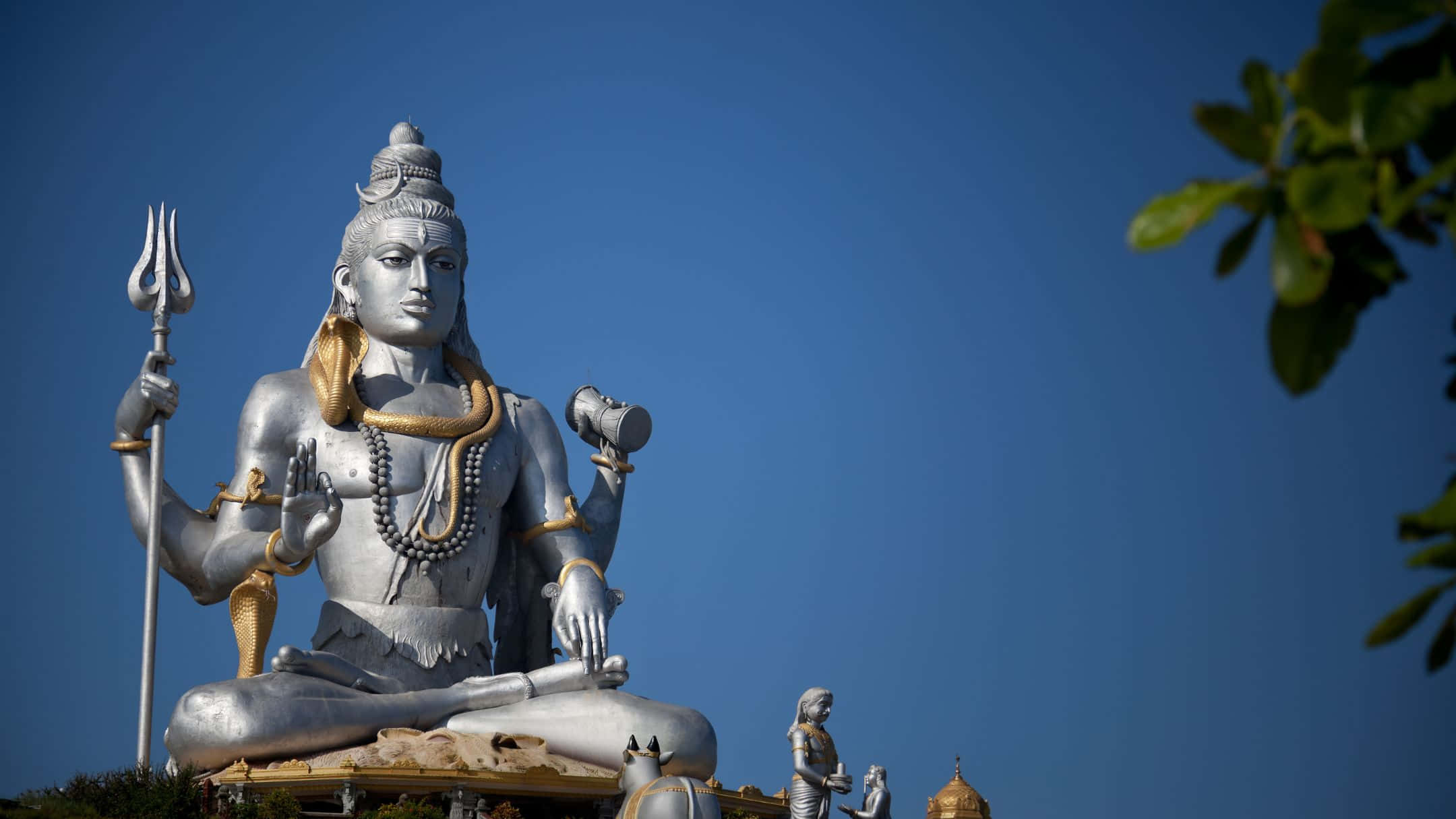 Shiva 桌布 壁紙 — 一座高聳的銀色濕婆神像，飾有金色裝飾，在綠葉環繞的清澈藍天下安詳地坐著。