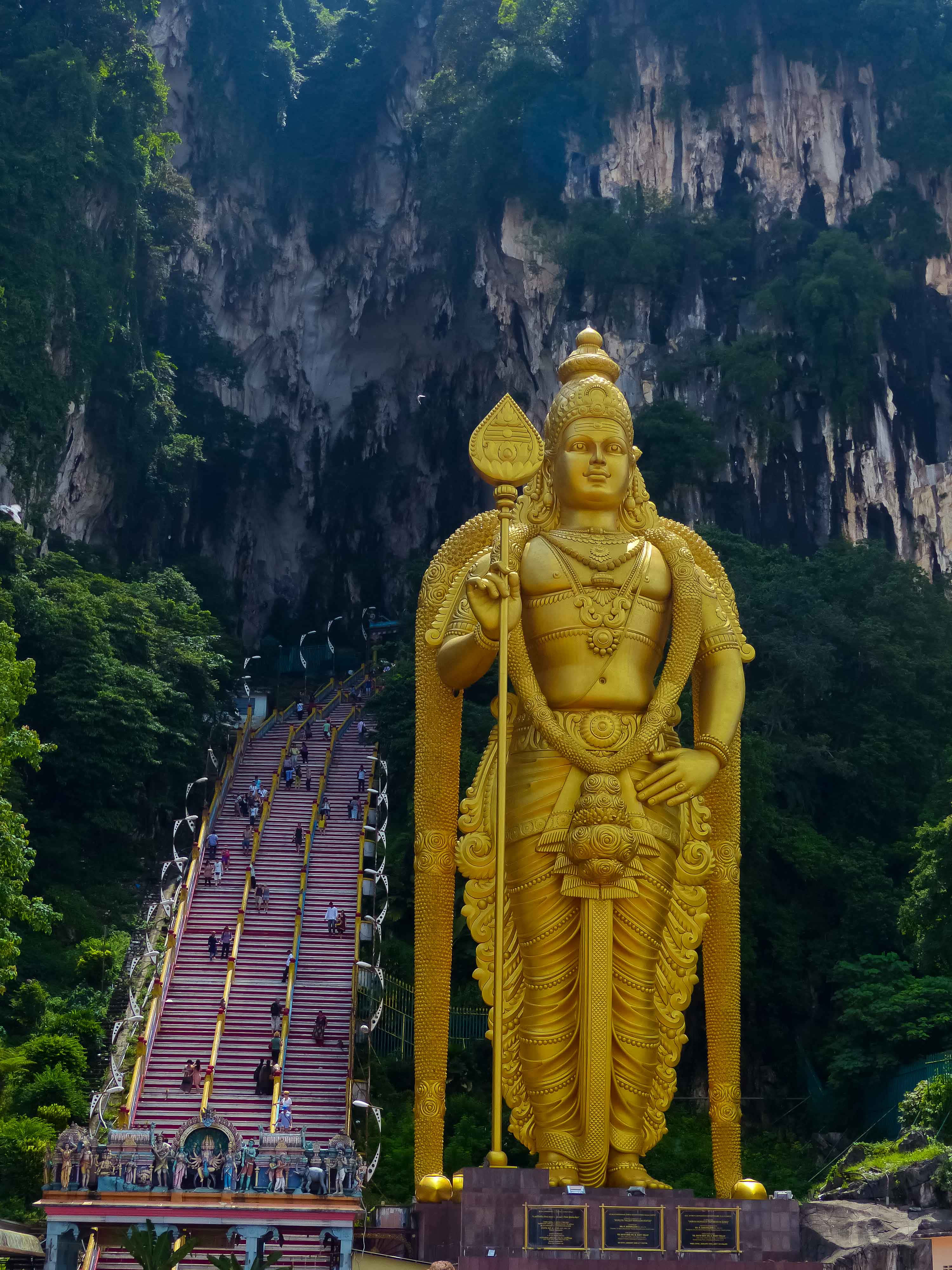 Murugan 桌布 壁紙 — 一座高聳的黃金穆魯干神像矗立在鬱鬱蔥蔥的綠色懸崖前，遊客正在攀登長長的樓梯。