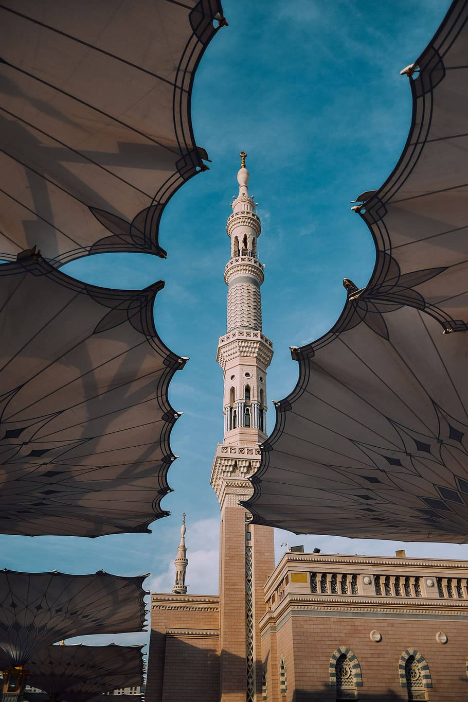 Madina 桌布 壁紙 — 一座高聳華麗的清真寺宣禮塔，穿過由大型圖案遮陽傘組成的幾何框架，映襯著清澈的藍天。