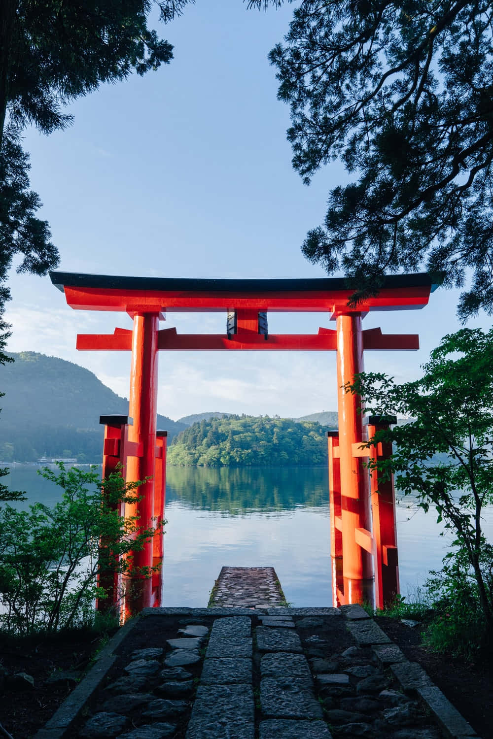 Shinto 桌布 壁紙 — 一座鮮紅色的鳥居矗立在寧靜的湖邊，四周是茂密的綠樹和遠山，天空清澈湛藍。
