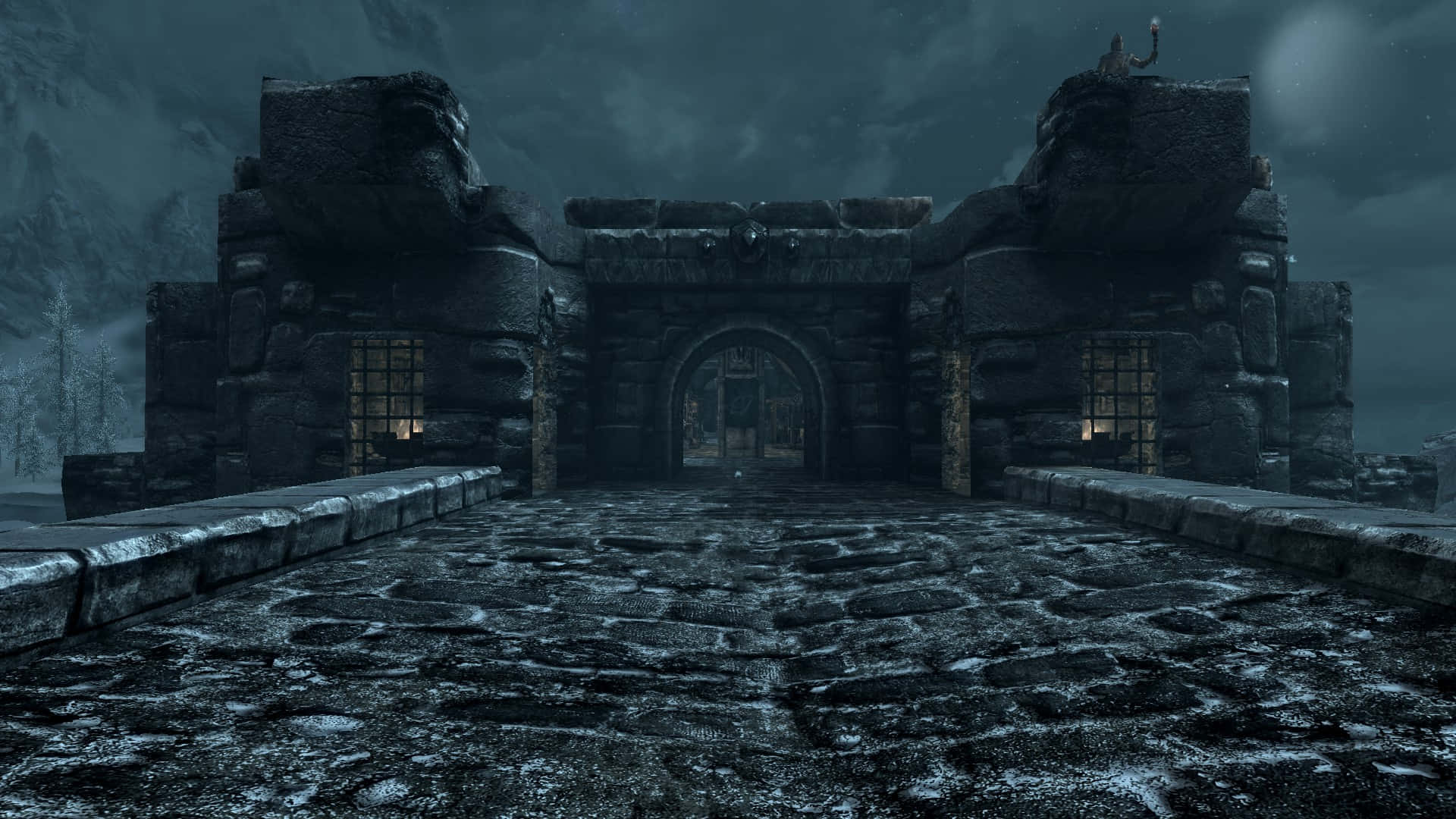 Skyrim Windhelm 桌布 壁紙 — 一座黑暗的石頭堡壘大門，帶有發光的窗戶，矗立在暴風雨夜空的雪地山景中。