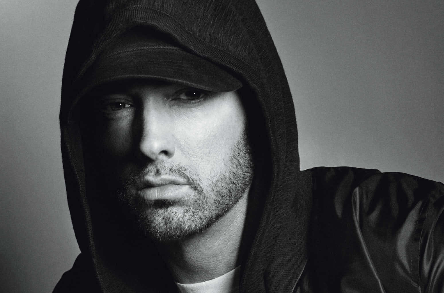 Eminem 桌布 壁紙 — 一張充滿情緒的黑白肖像照，捕捉一名身穿連帽外套的男子，在樸素灰色背景前展現強烈眼神。