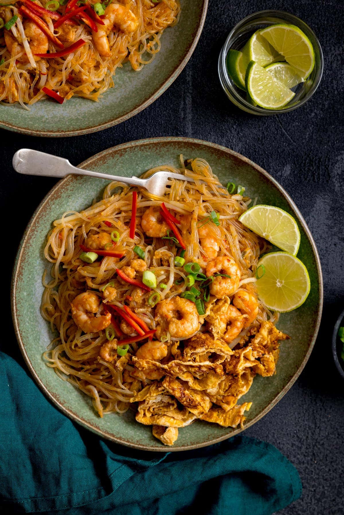 Noodles 桌布 壁紙 — 一張充滿活力的俯拍照片，兩碗蝦仁泰式炒河粉，飾以青檸角、紅辣椒和青蔥，置於深色紋理表面上。