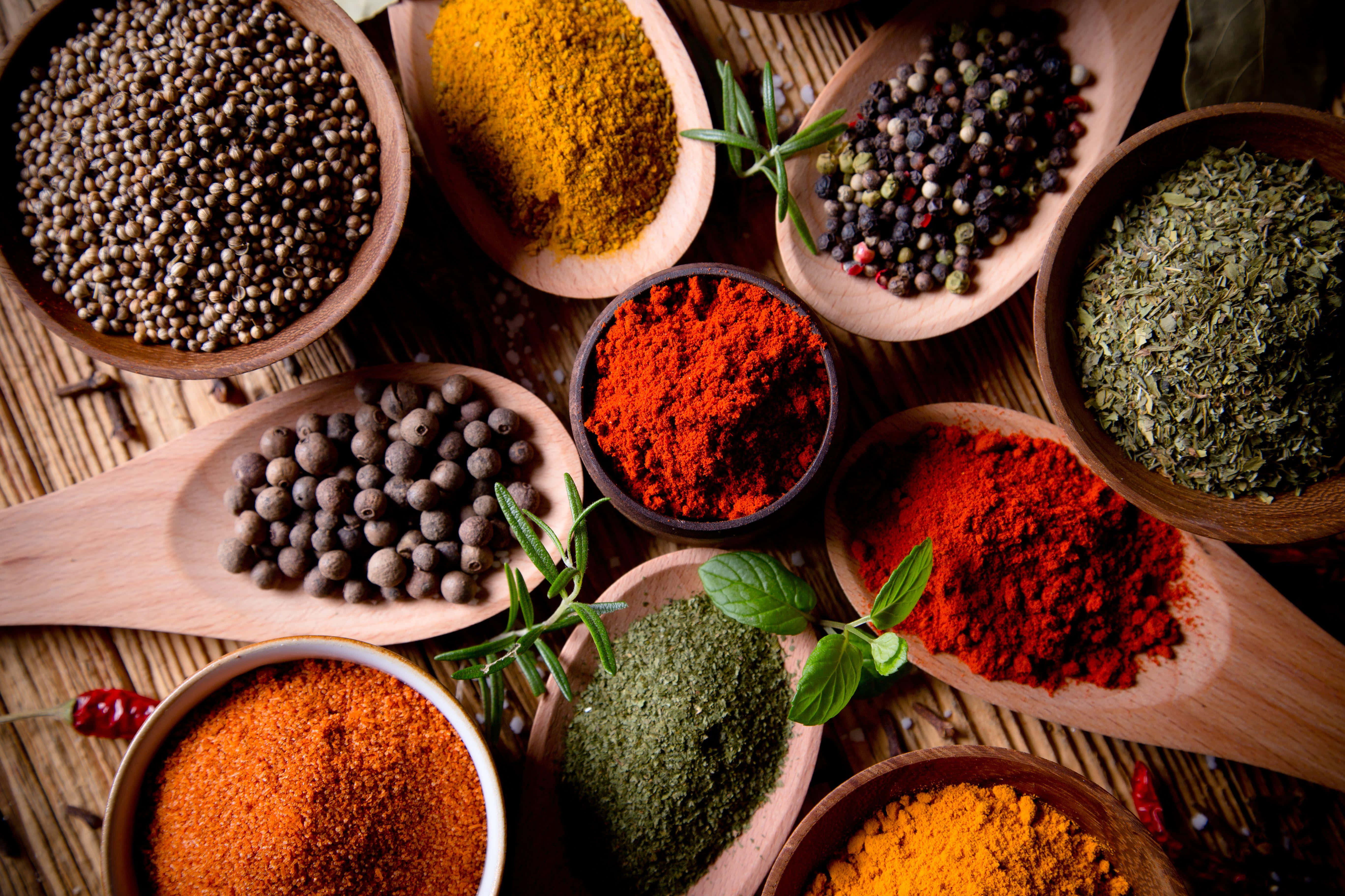 Spices 桌布 壁紙 — 一張充滿活力的俯拍照片，展示木勺和碗中各種香料和香草，擺放在編織墊上。