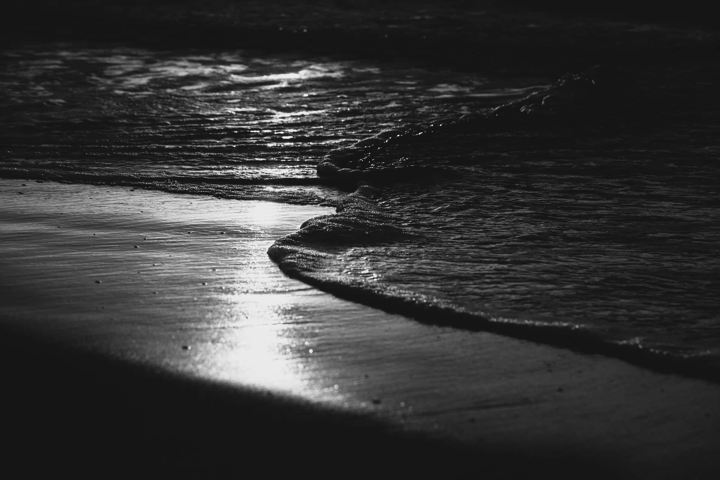 Black And White Beach 桌布 壁紙 — 一張單色拍攝，海浪輕輕湧上沙灘，濕沙上有明亮倒影。