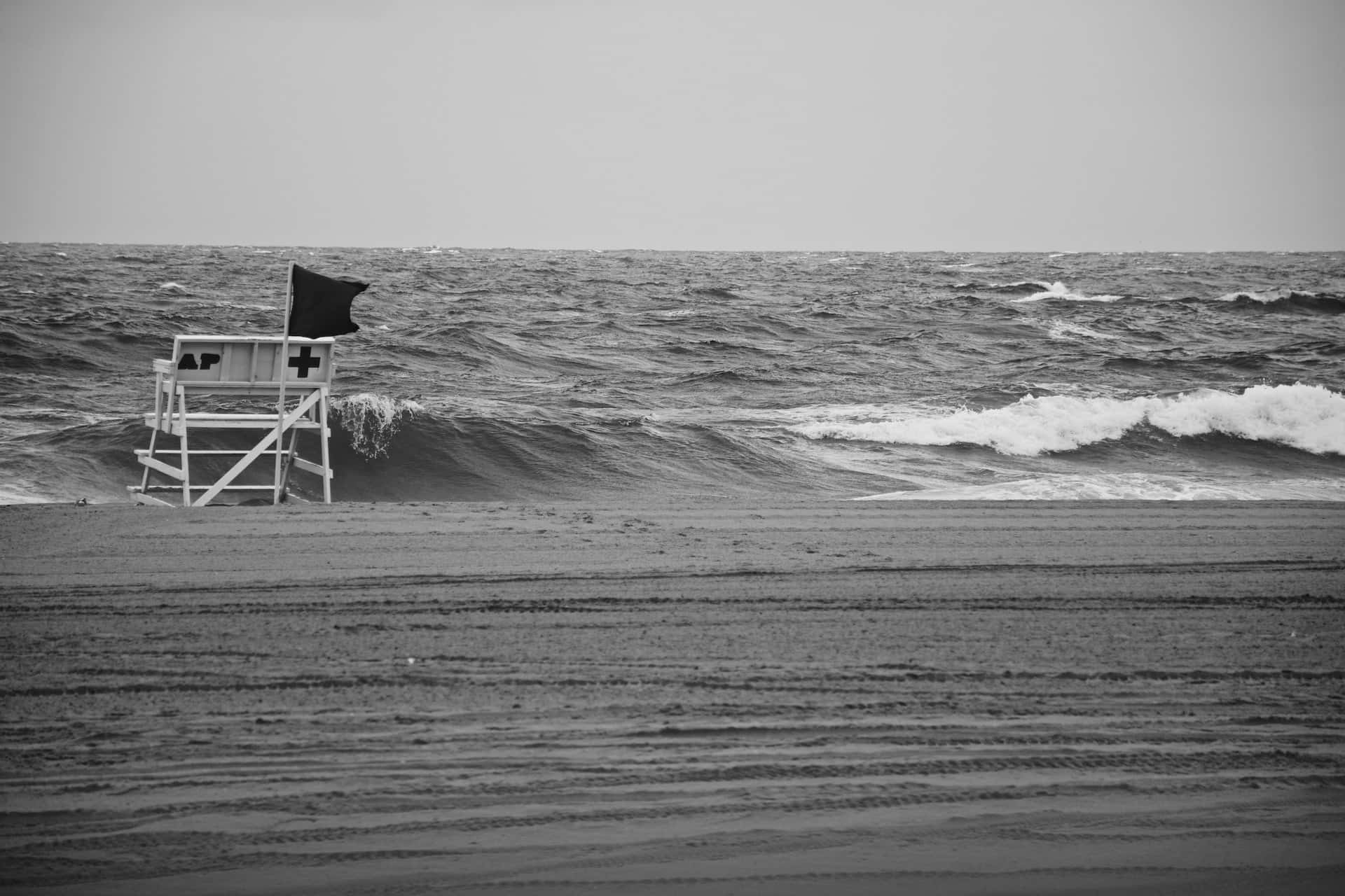 Black And White Ocean 桌布 壁紙 — 一張孤獨的救生員椅聳立在風吹沙灘上，黑暗的波浪在灰色天空下拍打著海岸。