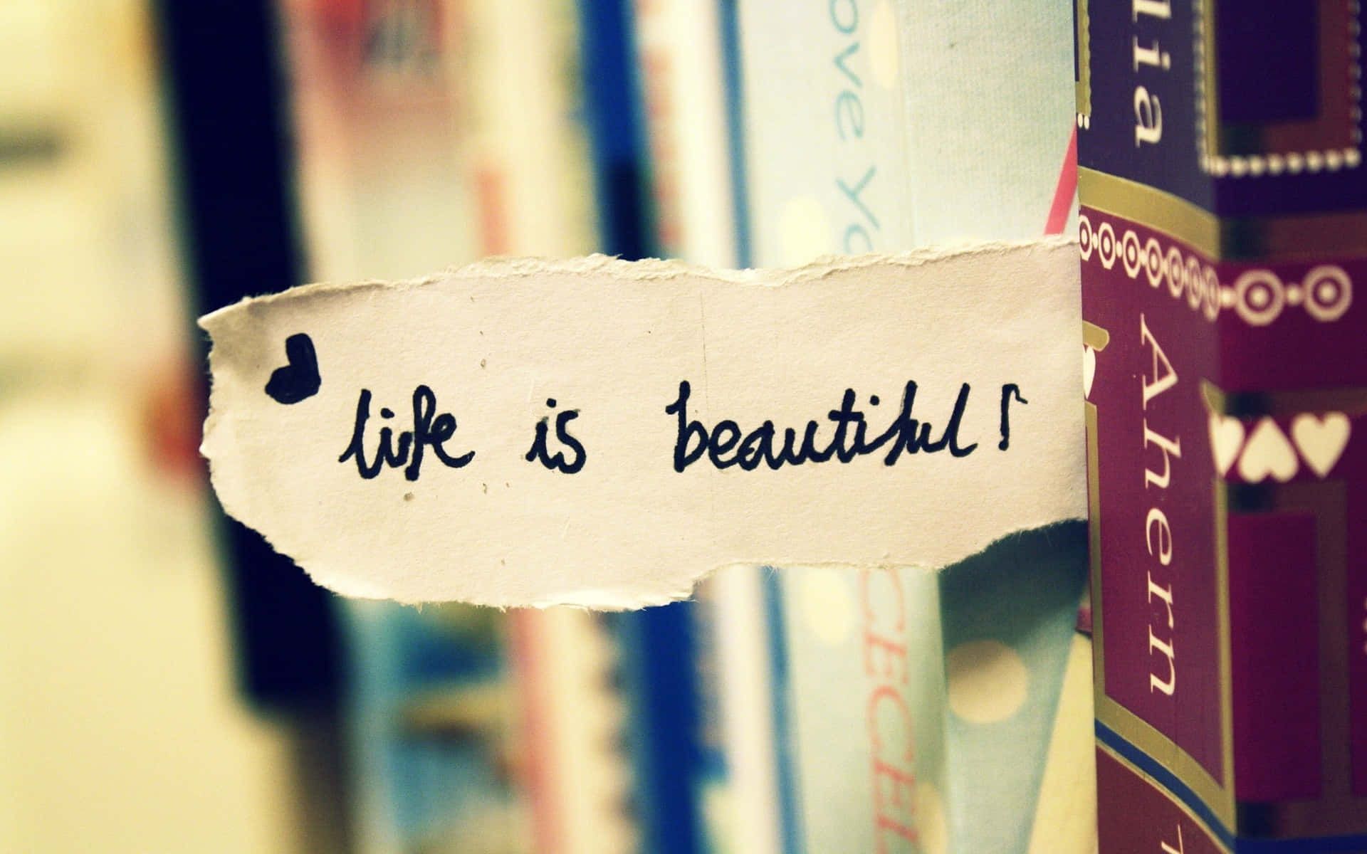 正面語錄 桌布 壁紙 — 一張寫著 life is beautiful! 並帶有心形圖案的破爛紙條，用膠紙貼在一個放滿彩色書籍的書架上。
