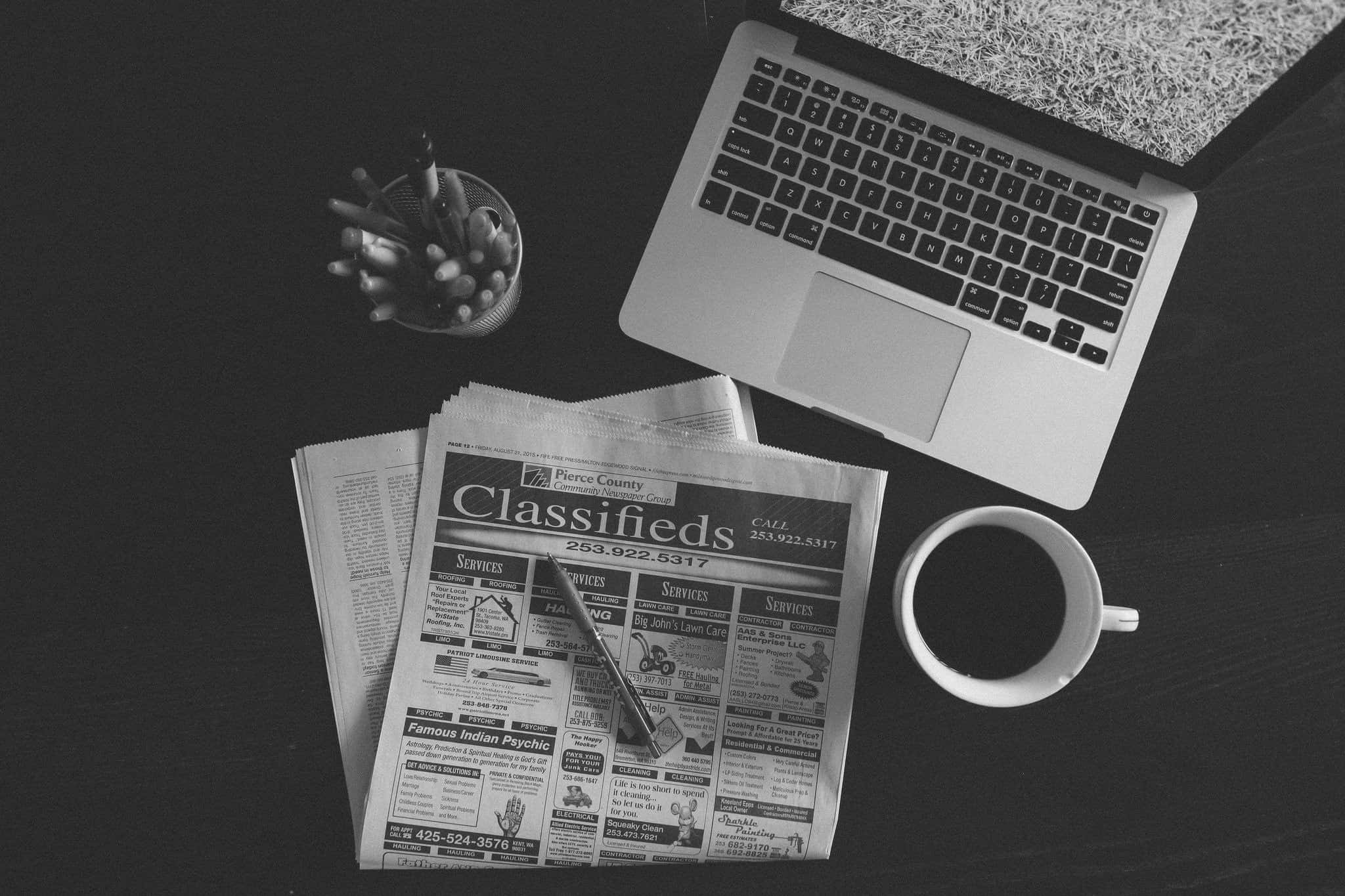 Black And White Newspaper 桌布 壁紙 — 一張從上方拍攝的單色照片，顯示深色書桌上的手提電腦、報紙分類廣告和咖啡杯。