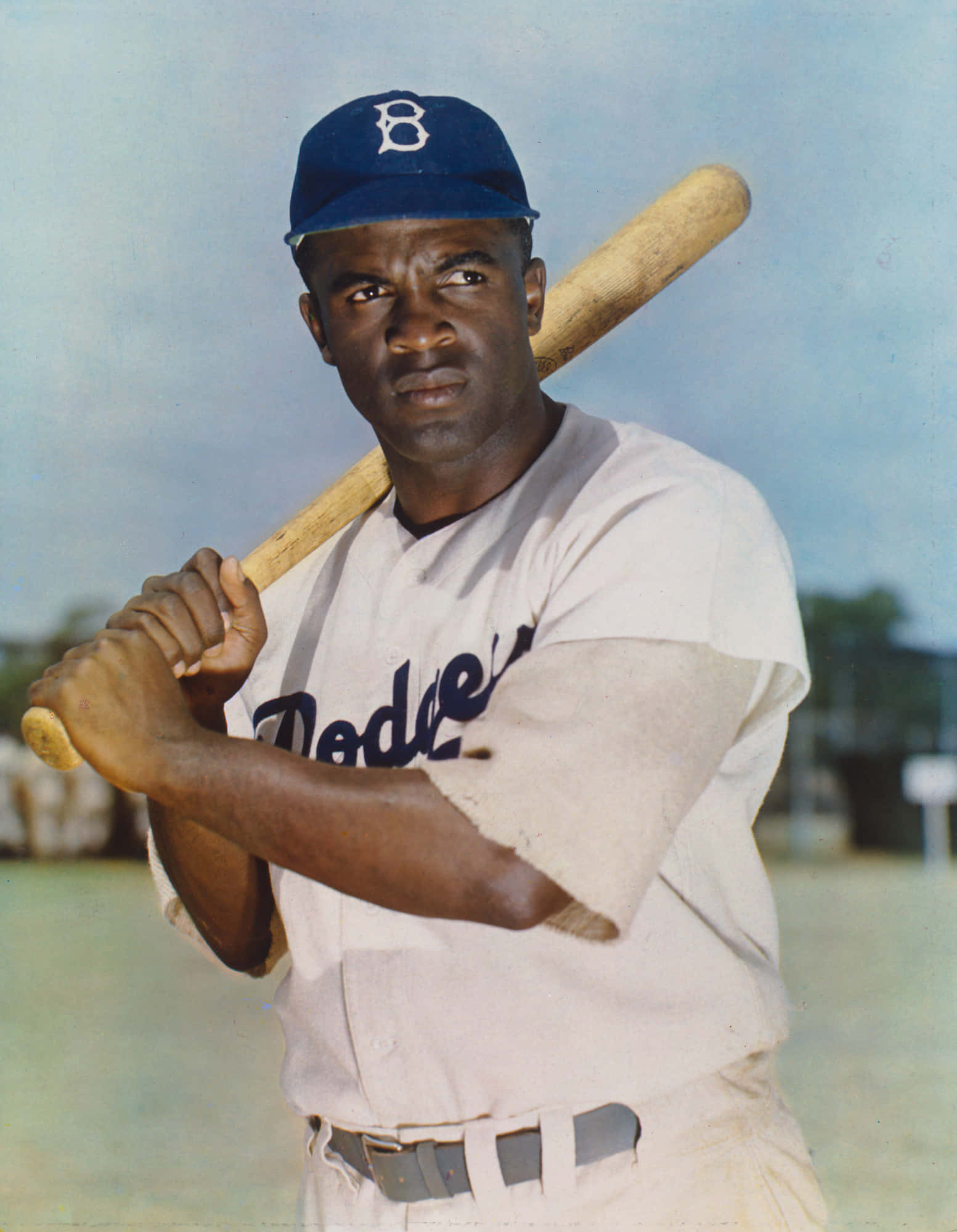 Jackie Robinson 桌布 壁紙 — 一張復古肖像，一名黑人棒球運動員身穿道奇隊制服，在柔和天空背景下專注地握著球棒。