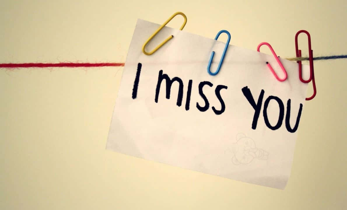 I Miss You 桌布 壁紙 — 一張手寫便條寫著「我想你」，掛在一條紅色繩子上，用彩色萬字夾夾著，背景是柔和的米色牆壁。