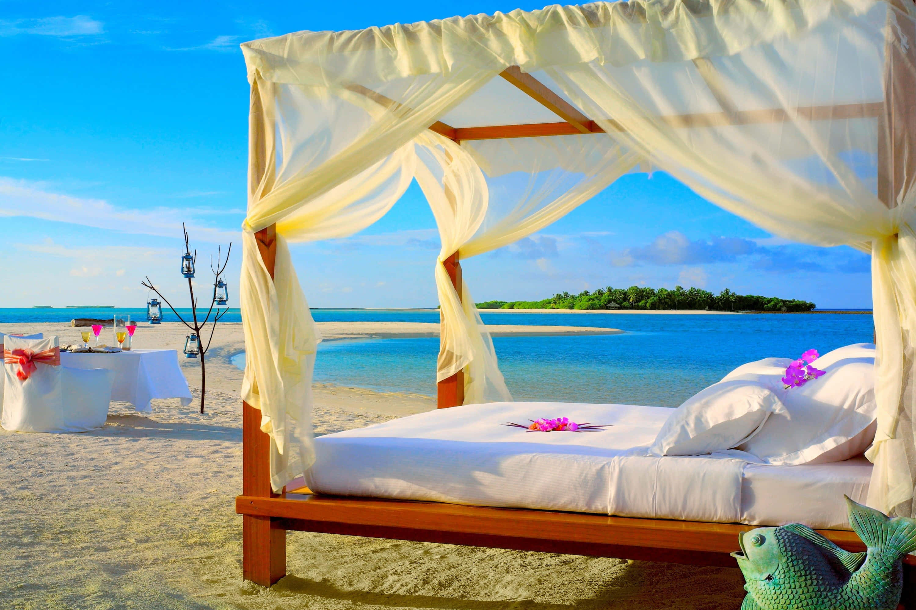 Beach Daybed 桌布 壁紙 — 一張木製躺椅，配有米色窗簾和粉紅色花朵，放置在沙灘上，俯瞰著碧綠的海水和遠處的綠色島嶼，天空湛藍明亮。