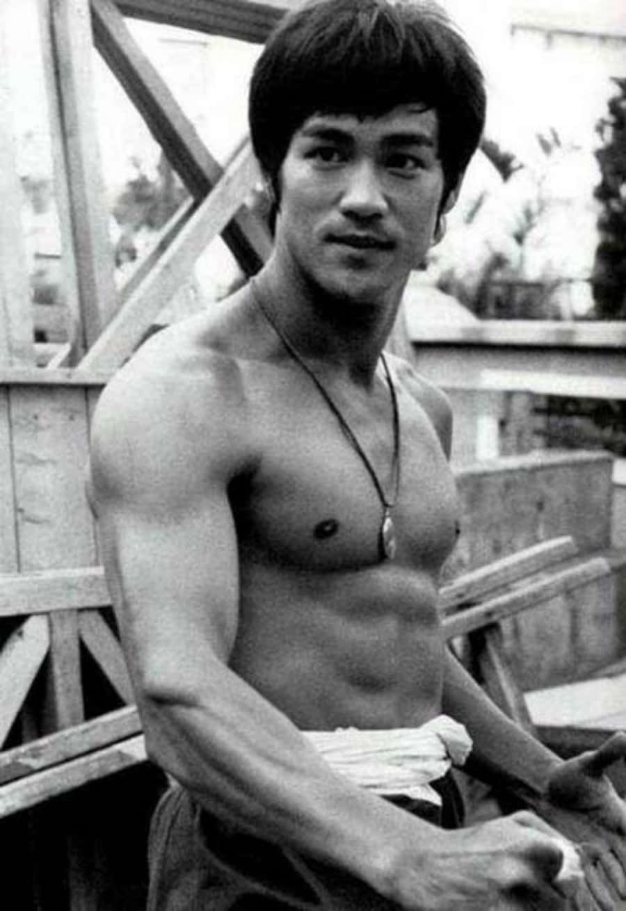 李小龍赤裸上身戴著項鍊坐在戶外木結構附近。