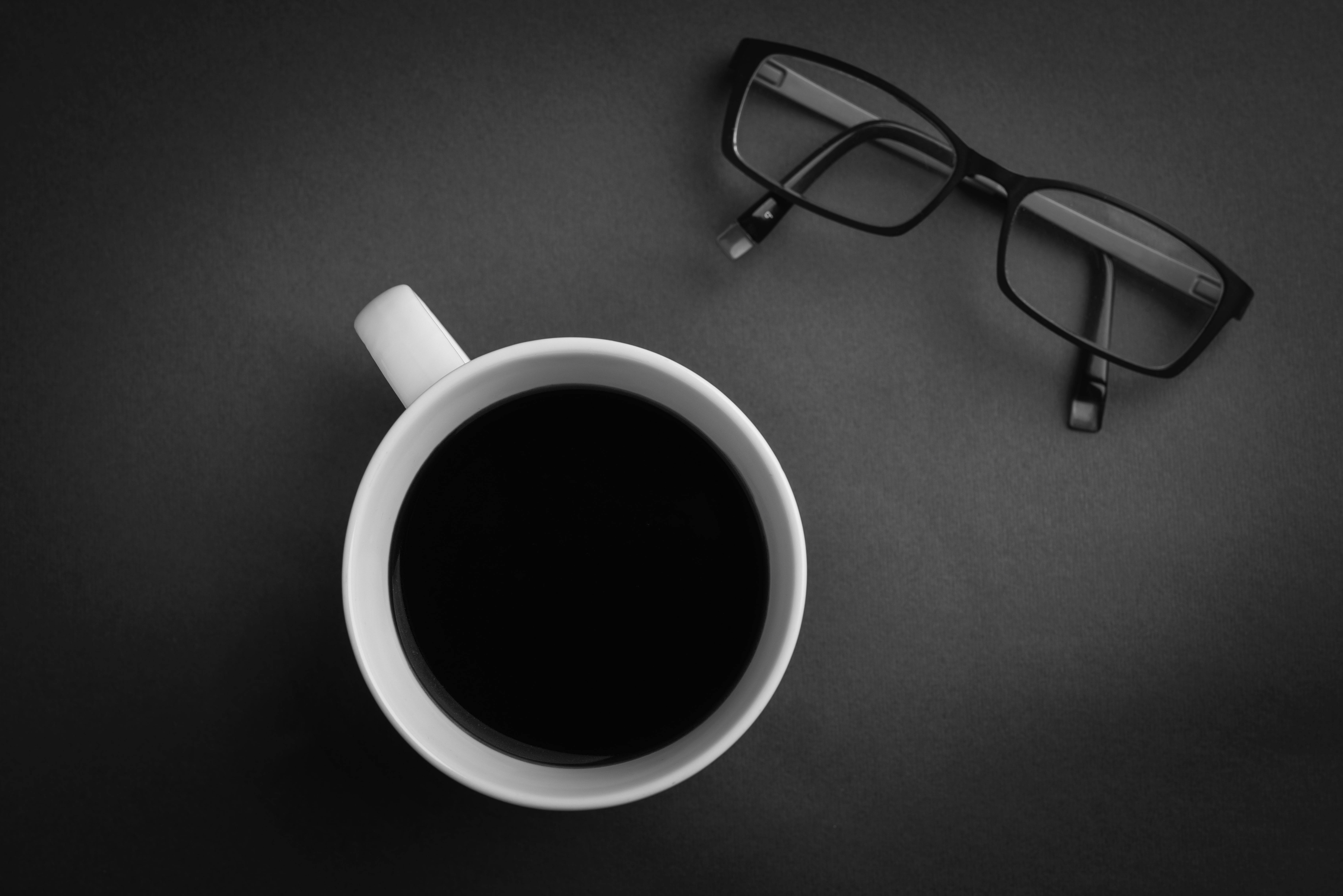 Coffee Aesthetic 桌布 壁紙 — 一張極簡黑白俯拍照片，展示白色咖啡杯和老花眼鏡放在深色紋理表面上。