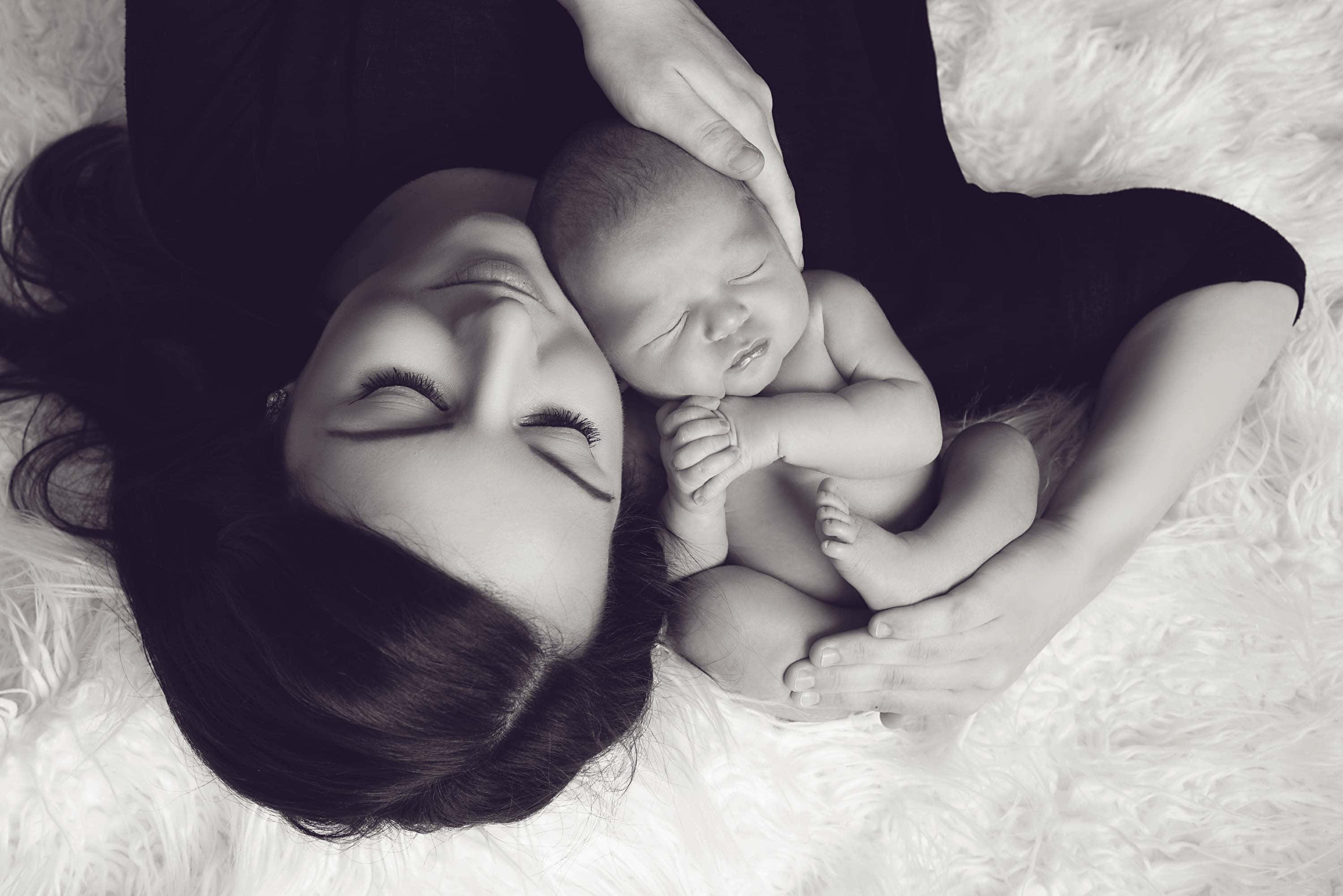 Newborn Baby 桌布 壁紙 — 一張溫柔的黑白肖像，母親在 fluffy 白色地毯上懷抱著熟睡的新生兒。