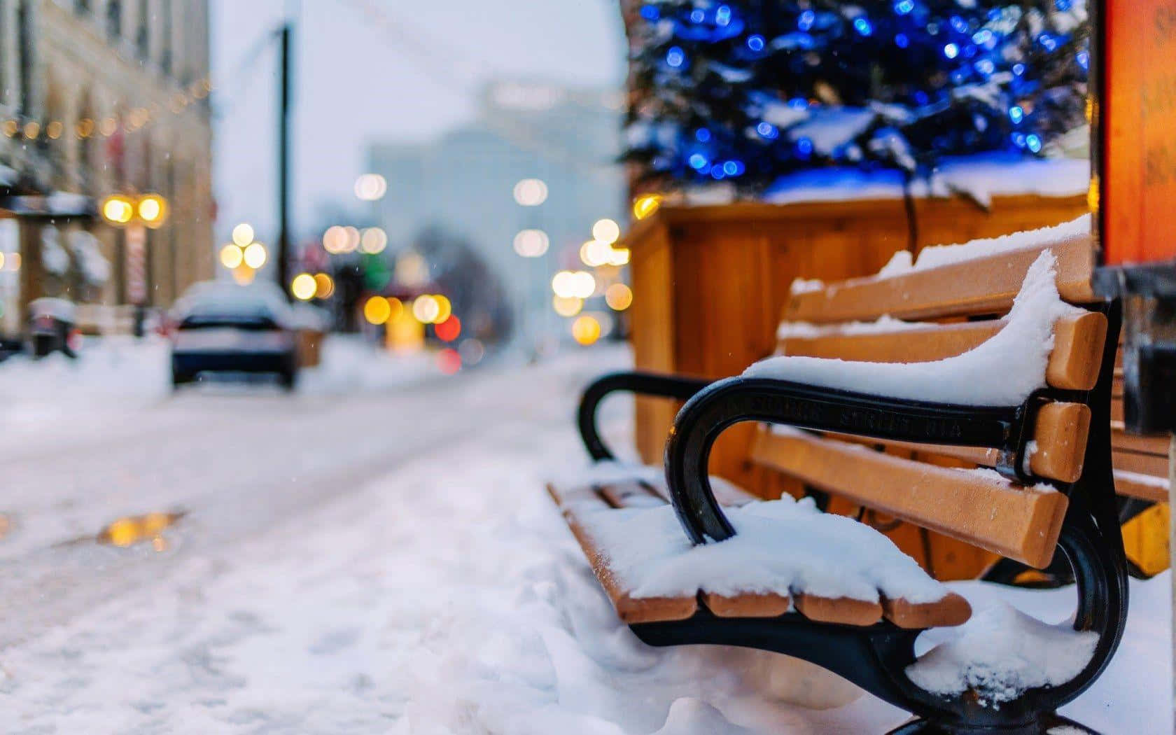 Winter Cityscape 桌布 壁紙 — 一張灑滿雪花的木長凳靜靜地放在冬日城市街道上，背景中有街燈和裝飾樹散發的發光散景燈光。