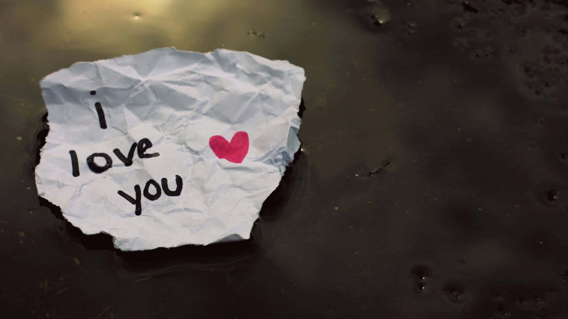 我愛你 桌布 壁紙 — 一張皺巴巴的白色便條，上面寫著「i love you」並帶有一個粉紅色心形，在溫暖燈光下漂浮在潮濕的深色路面上。