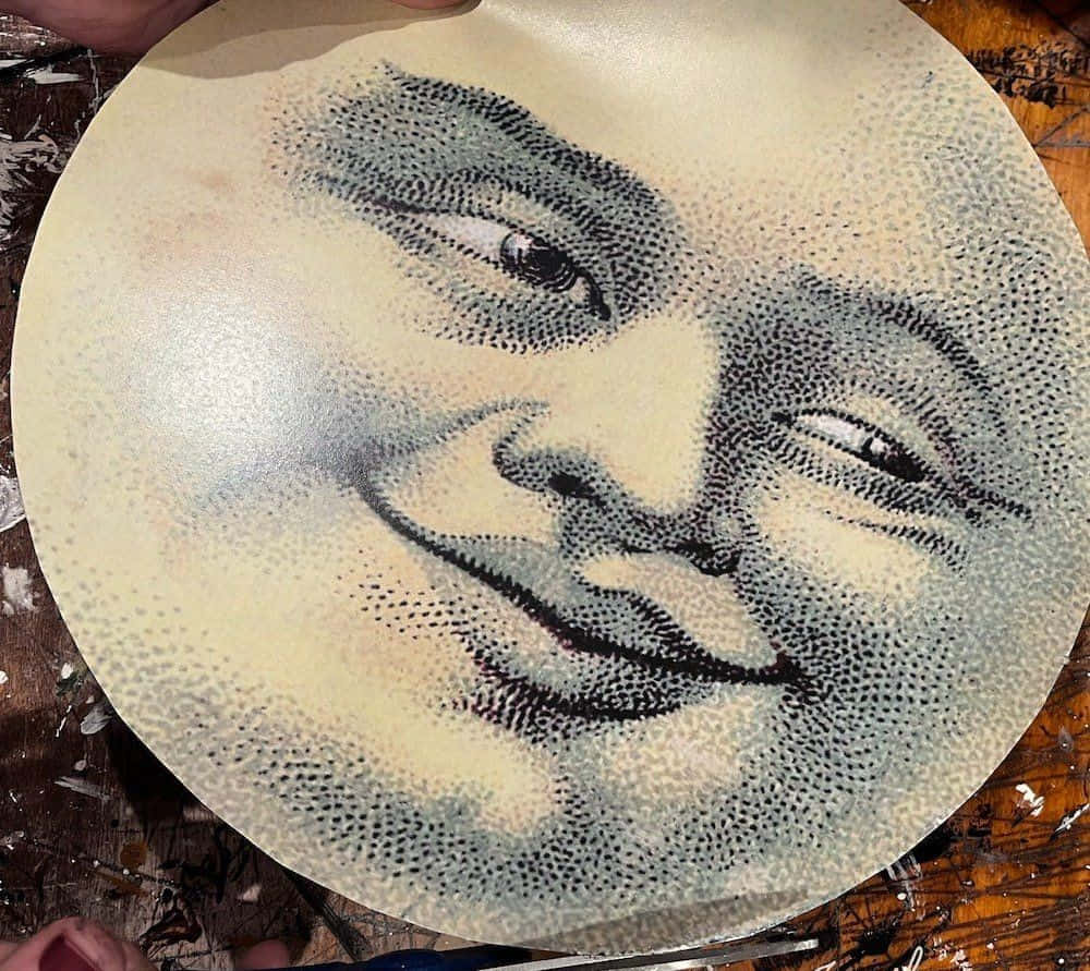 一張神秘月面的抽象圖像 壁紙 — 一個圓形點陣肖像，描繪一個微笑、閉著眼睛的臉，位於一個有紋理的表面上，由雙手捧著。