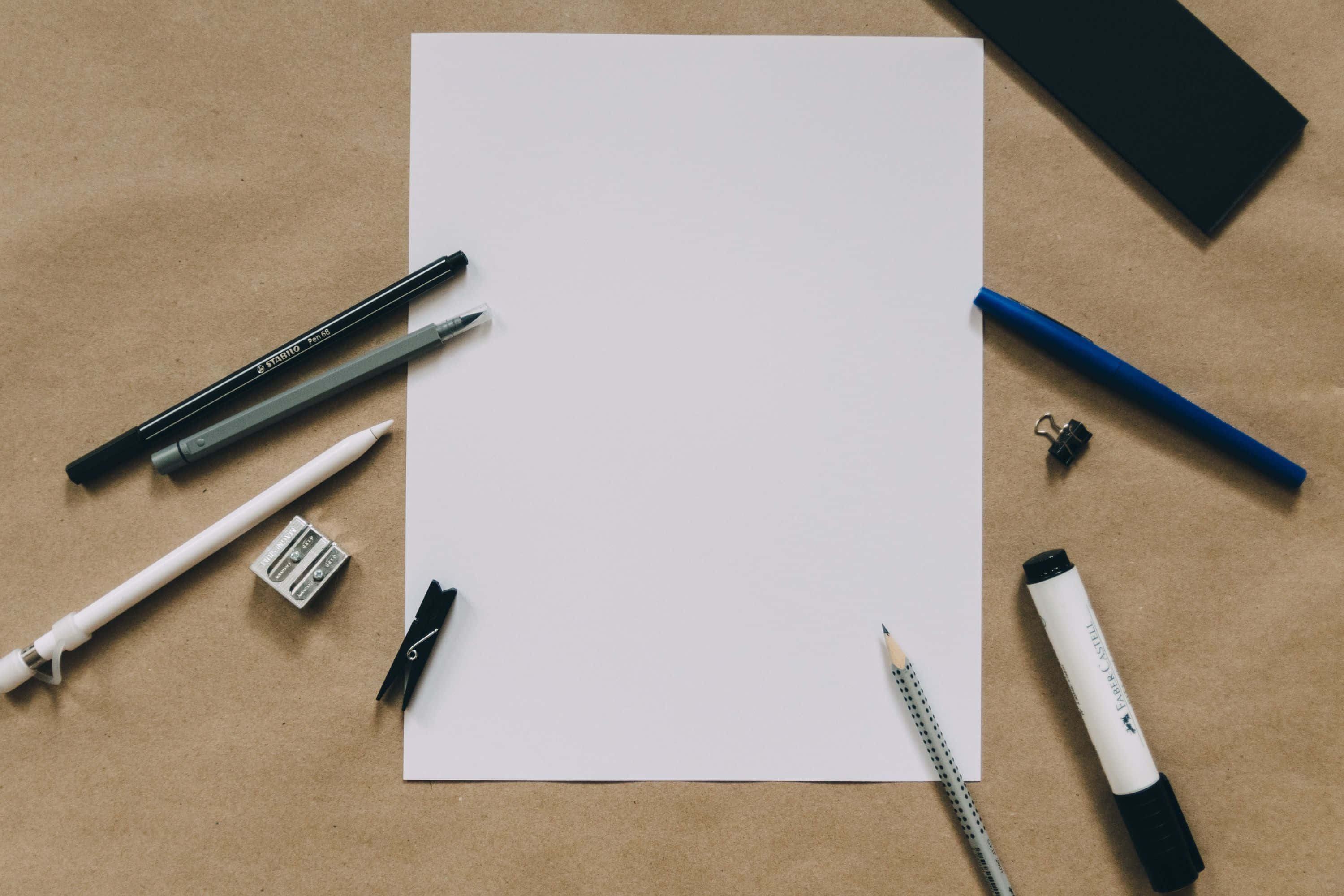 paper 桌布 壁紙 — 一張空白白紙，周圍環繞著原子筆、鉛筆和文具，放在棕色紙上。