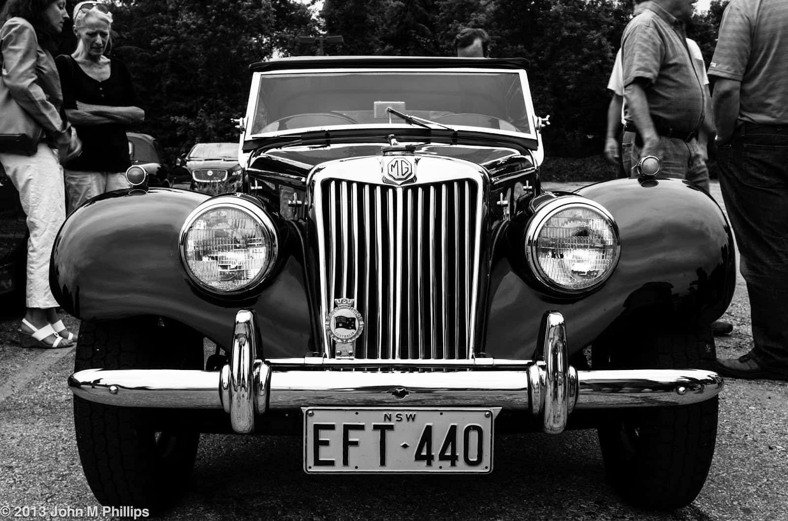 Black And White Vintage Car 桌布 壁紙 — 一張經典黑白照片，展示一輛配備鍍鉻格柵和圓形頭燈的復古MG開篷車，在戶外聚會中被圍觀者包圍。