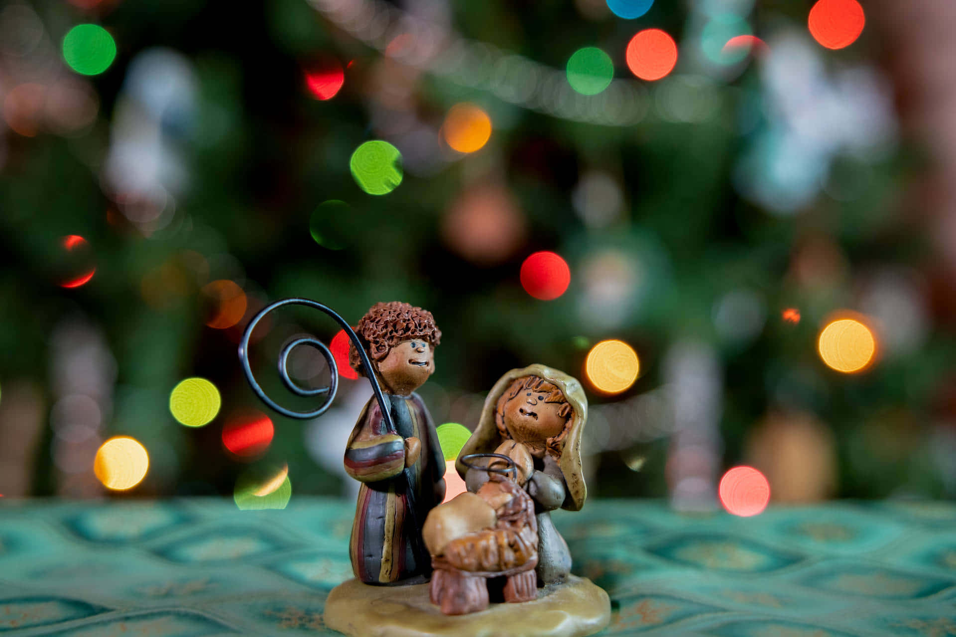 Nativity Scene 桌布 壁紙 — 一張聖誕馬槽雕像的特寫照片，有牧羊人和瑪利亞懷抱嬰兒耶穌，背景是色彩繽紛的聖誕燈飾散景。