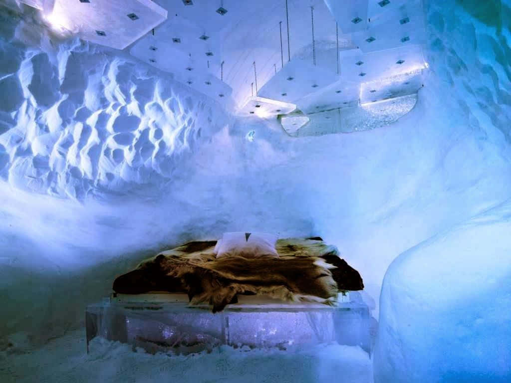 Ice Hotel 桌布 壁紙 — 一張舒適的床鋪著毛毯，位於發光的藍色冰洞中，牆壁有紋理，天花板有幾何燈光。