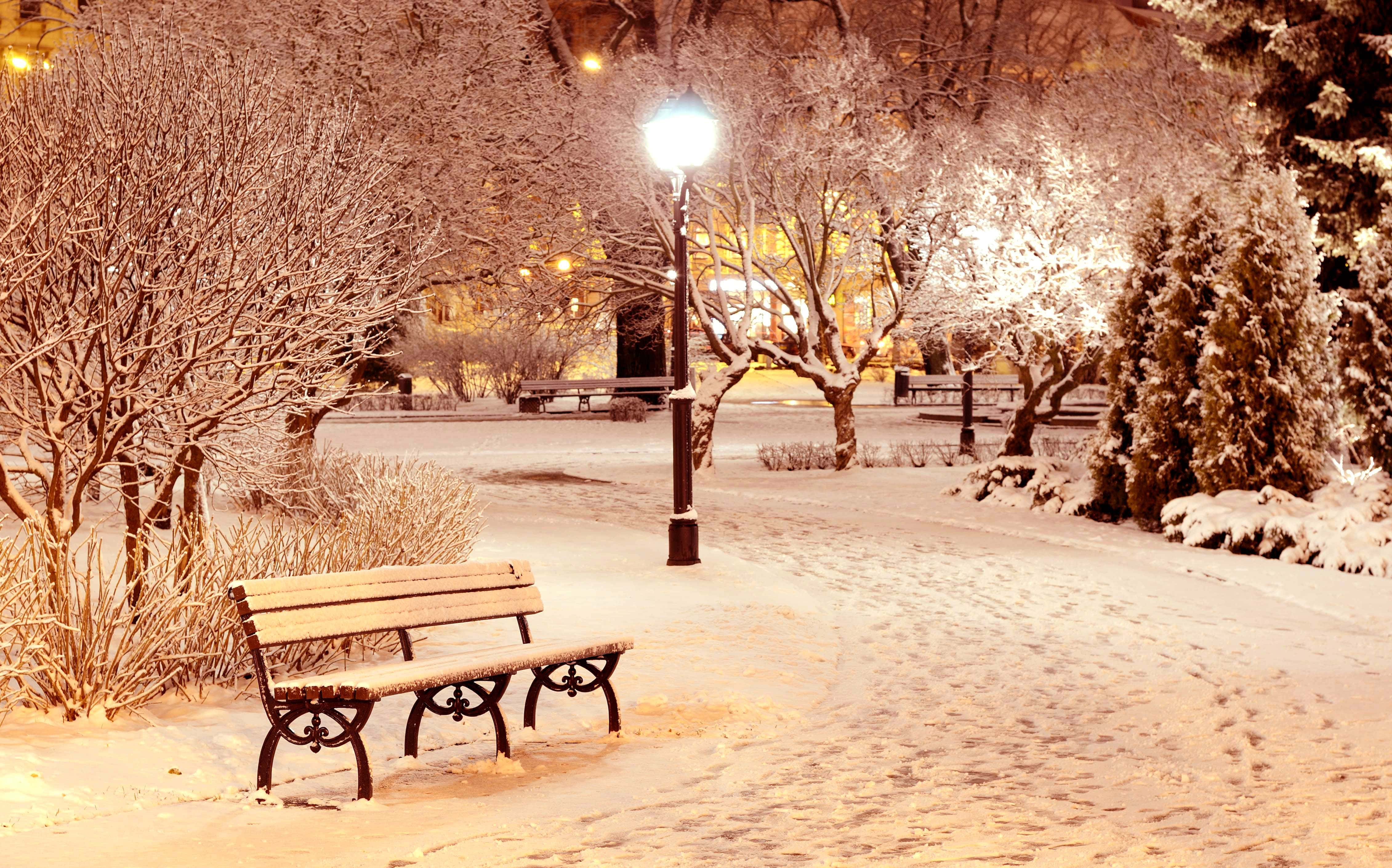 冬季 桌布 壁紙 — 一張被雪覆蓋的公園長椅在夜晚的路燈下溫暖地發光，腳印蜿蜒穿過白色的景觀。